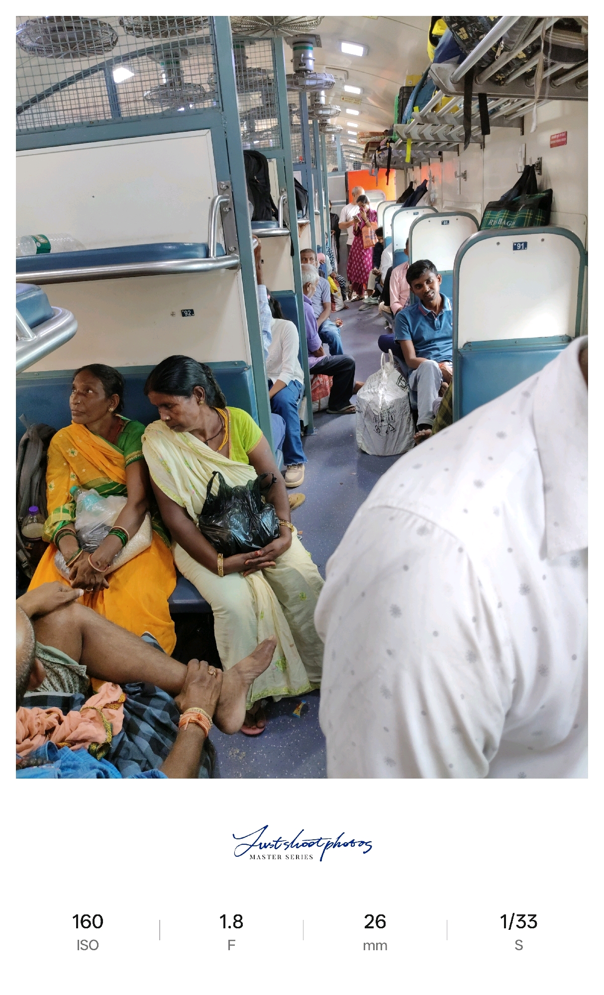 The image depicts the interior of a crowded train carriage in Chhapra, India. Several passengers are seated on blue benches, with luggage visible on overhead racks and on the floor. In the foreground, two women are seated, one wearing a yellow and green sari, and the other a cream-colored sari. The woman on the right holds a black plastic bag. To their left, a person's legs are visible, partially covered by a patterned cloth. Other passengers, including men in the background, are seen sitting and standing. The carriage features blue and white paneling and overhead fans. A white shopping bag with a print is placed on the floor between seats. The overall scene is a snapshot of daily life on a train, characterized by close proximity and visible belongings.