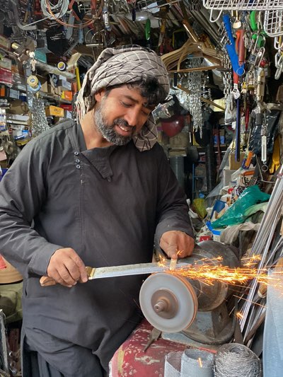 A man with a head covering is sharpening a blade on a rotating grinder, producing sparks. The setting appears to be a crowded workshop or store in Kandahar, Afghanistan, with various tools and hardware items visible in the background. The man is wearing a dark-colored garment and has a beard. He is focused on the task at hand, holding the blade against the grinder.