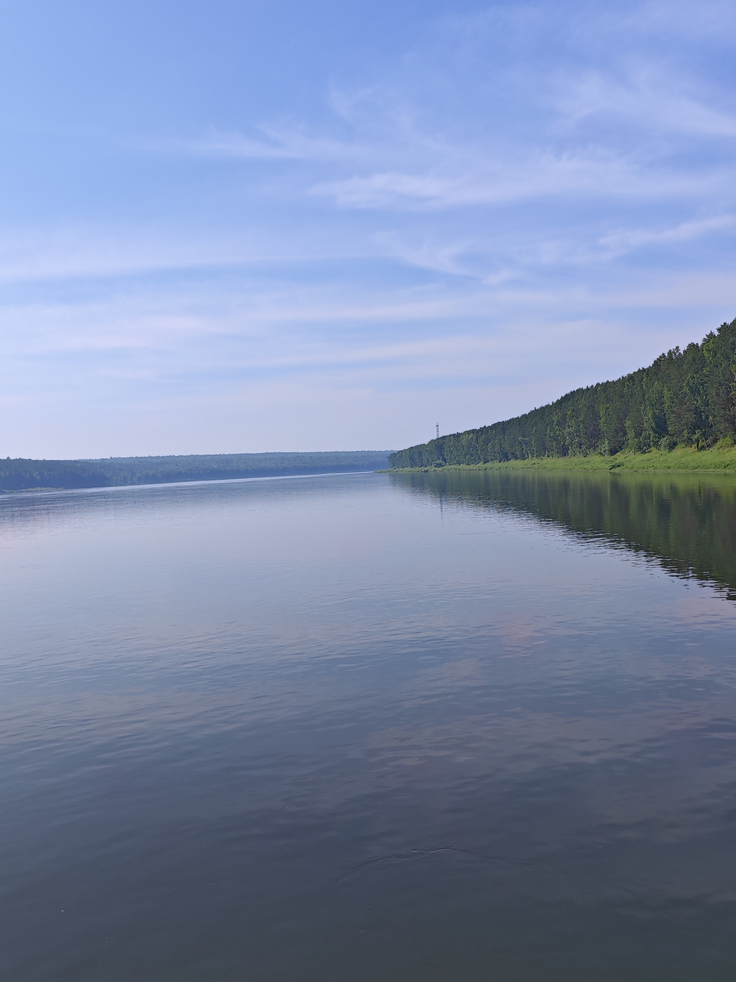 The image shows a wide, calm river with a forest lining the far bank. The sky is blue with scattered white clouds. A transmission tower is visible on the left side of the forest. The scene is likely in the city of Piskunovka, Russia, given the mention of its location in the prompt. There are no people or animals visible in the image. The water reflects the trees and sky, creating a serene natural landscape.