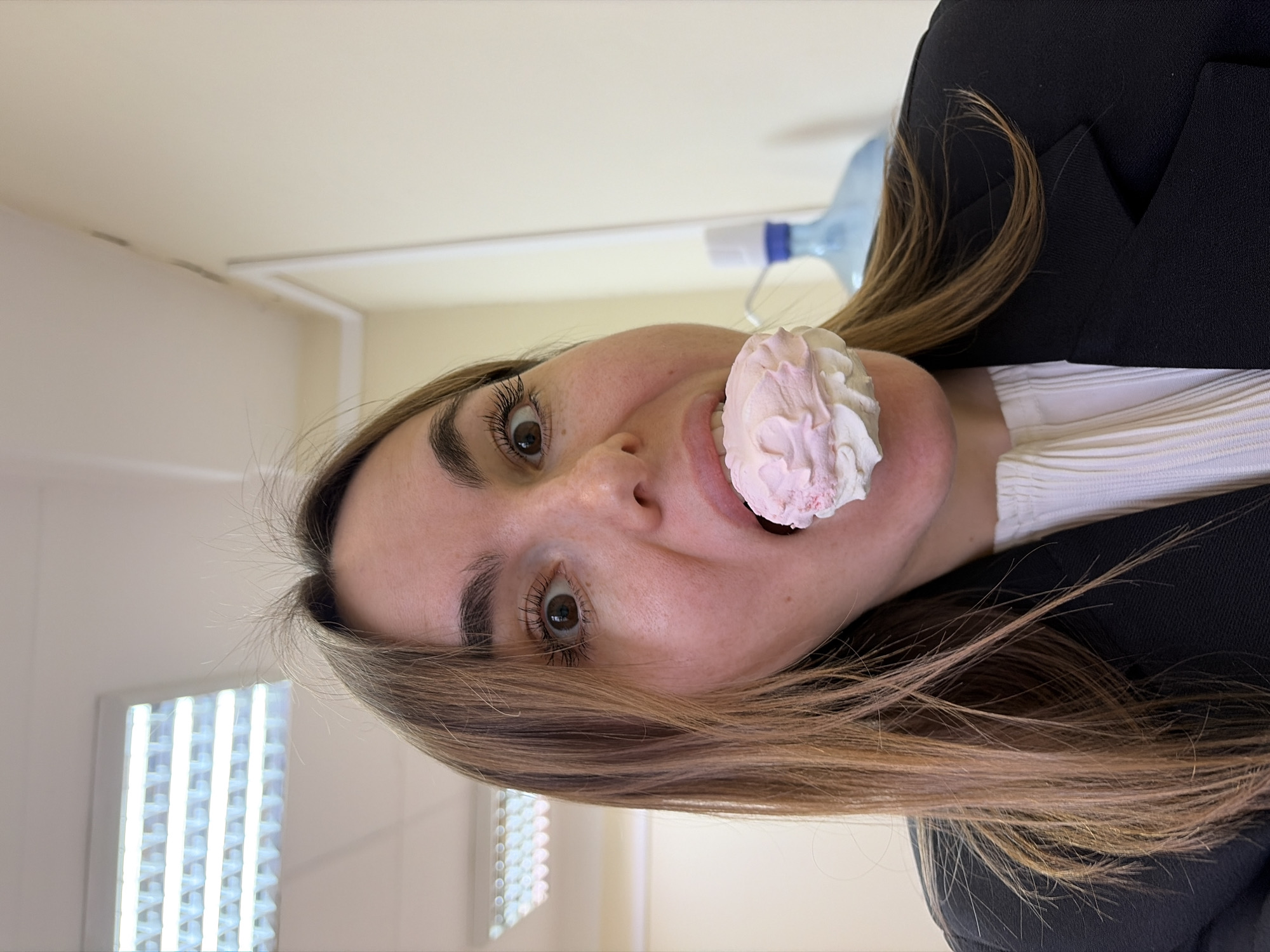 The image depicts a woman indoors. She has brown hair, brown eyes, and fair skin. She is wearing a black blazer over a white, pleated top. She is holding a pink and white frosted treat in her mouth. In the background, there's a fluorescent light fixture, a beige wall, and a blue water bottle. There is no verifiable evidence to identify the location as Izhevsk, Russia.