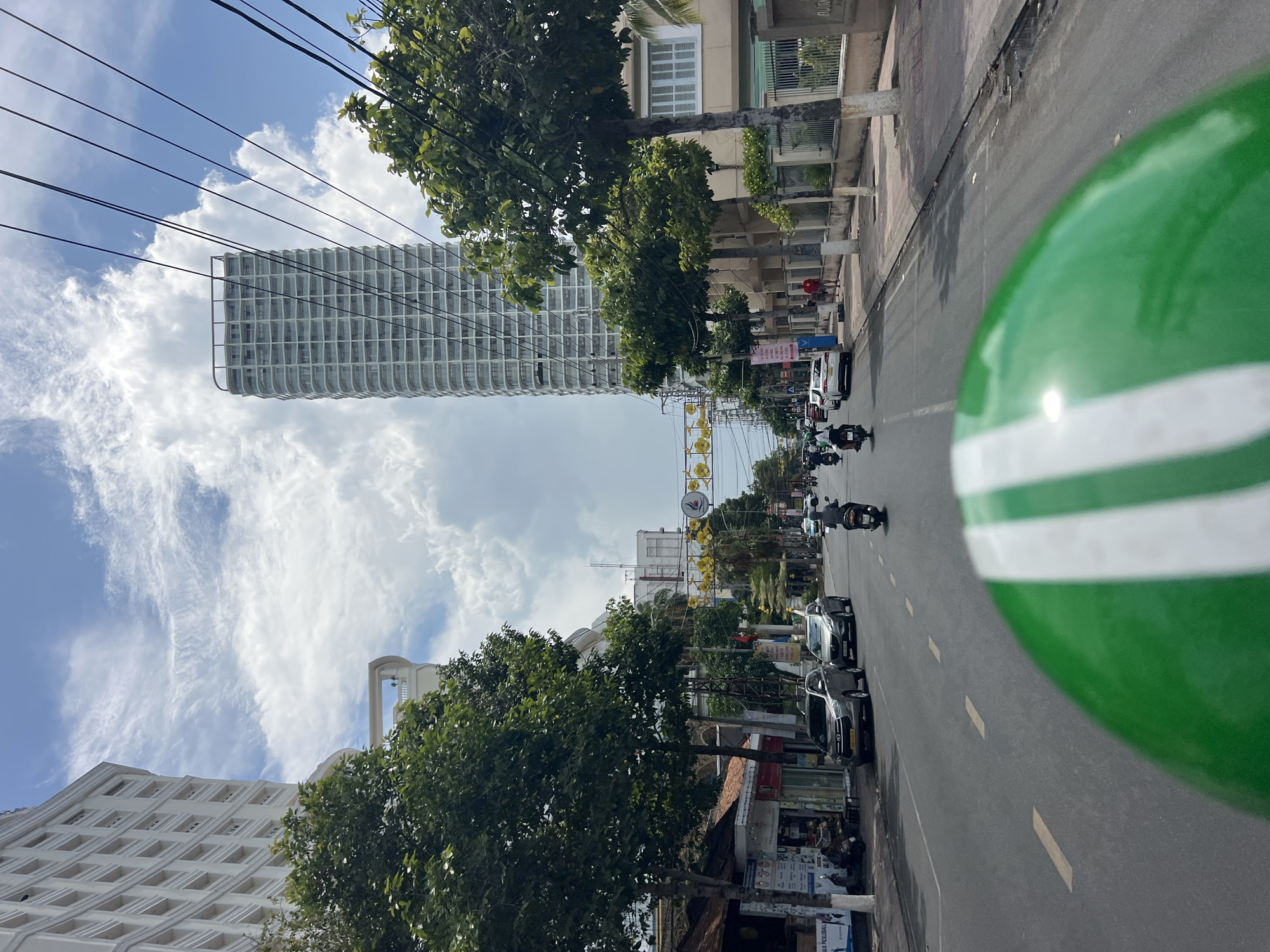 The image captures a street scene in Nha Trang Ward, Vietnam, looking down a road lined with buildings and trees. A modern, high-rise building with a grid-like facade stands tall on the left side of the street. Utility wires stretch across the sky.
The road is filled with motorcycles and cars. A green and white helmet is visible in the foreground, partially obscuring the view, suggesting the image was taken from a vehicle. Buildings line the street, some with shops and businesses. Trees provide shade along the sidewalks. The sky is partly cloudy.