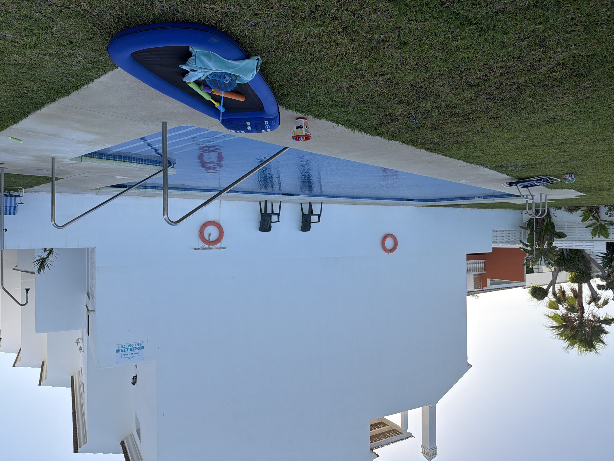 The image depicts a residential complex with a swimming pool in Estepona, Spain. The main building, a white stucco structure, occupies the majority of the frame. To the left, a palm tree and other lush greenery are visible, along with parts of neighboring orange and white buildings. The swimming pool, filled with clear blue water and featuring a tiled bottom, is situated in the foreground, bordered by a concrete deck. Two black plastic chairs are placed on the deck behind the pool. Two orange life preservers are mounted on the white wall of the building, one to the left and one to the right, each attached to a metal hook. Near the pool's edge, on the grass, lies a blue inflatable raft with various pool toys, including a green net and a yellow and orange flotation device. A small bucket with a cartoon character design sits on the concrete next to the raft. A lounge chair and a colorful ball are also present on the grass near the pool. The sky is clear and light blue, indicating daytime. A "For Sale" sign is affixed to the building's wall.