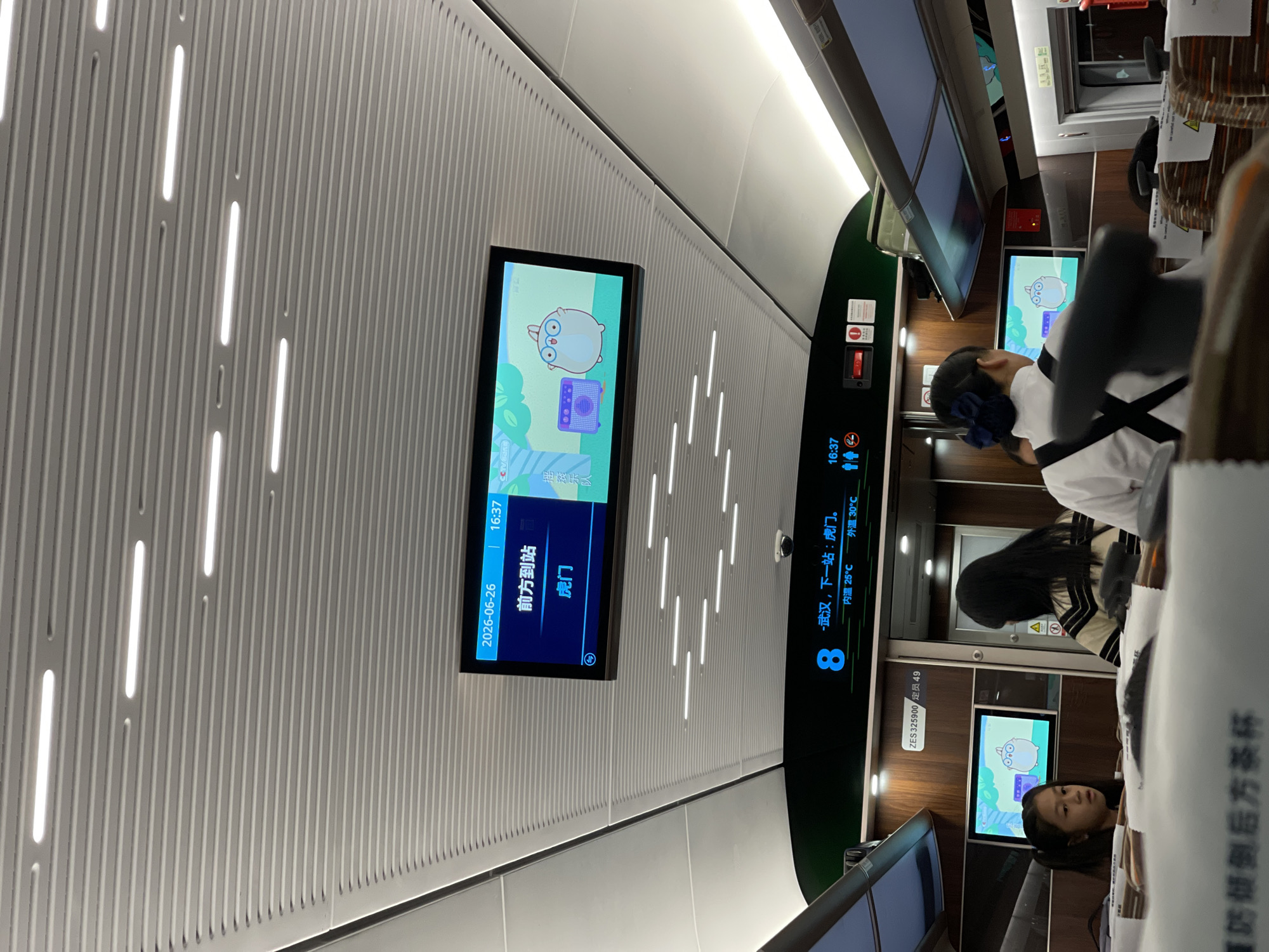The image shows the interior of a train carriage, likely on a high-speed rail line in China, specifically noted as Longhua District. The scene is oriented vertically, suggesting the photo was taken with the camera held in portrait mode.
The primary subjects are several passengers, including a young girl with dark hair and a blue hair accessory, seated and looking towards a screen. Another passenger with dark hair, wearing a striped top and a white collared shirt, is partially visible, also looking towards a screen. The train carriage is characterized by a modern, clean design with ribbed white walls and integrated LED lighting strips.
Several digital displays are visible. One prominent screen shows a cartoon character, likely a game or entertainment interface, with Chinese text. Another screen displays train information, including the date "2026-06-26", time "16:37", the current station "前方到站" (approaching station), and the next station "虎门" (Humen). A larger display panel on the right side shows the train number "8" and indicates "武汉,下一站:虎门" (Wuhan, next stop: Humen), along with interior and exterior temperatures: "内温25℃" (indoor temperature 25°C) and "外温30℃" (outdoor temperature 30°C). Another visible text reads "ZES 325900 定员49", which might be related to the train's model or capacity.
A red emergency button and some other warning signs are also present on the wall. The overall atmosphere appears calm, with passengers engaged with their screens or resting. The time indicated is late afternoon, and the exterior temperature suggests a warm day. The text "臺奶倾倒后方茶杯" (Milk to be poured behind the teacup) is visible on a white surface, possibly a warning or instruction related to food and drinks.