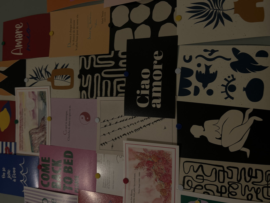 The image shows a collection of cards pinned to a wall, creating a mood board. The cards feature various designs and texts, including the phrase "Amore mio" in red, "Ciao Amore" in black and white, "Come Back to Bed" in pink, and "On se jette à l'eau" in blue. Other cards have inspirational quotes in French, signed by Mark Twain and Victor Hugo. There are cards displaying nature scenes, abstract shapes, and an illustration of a nude figure. Pins of different colors secure the cards in place.