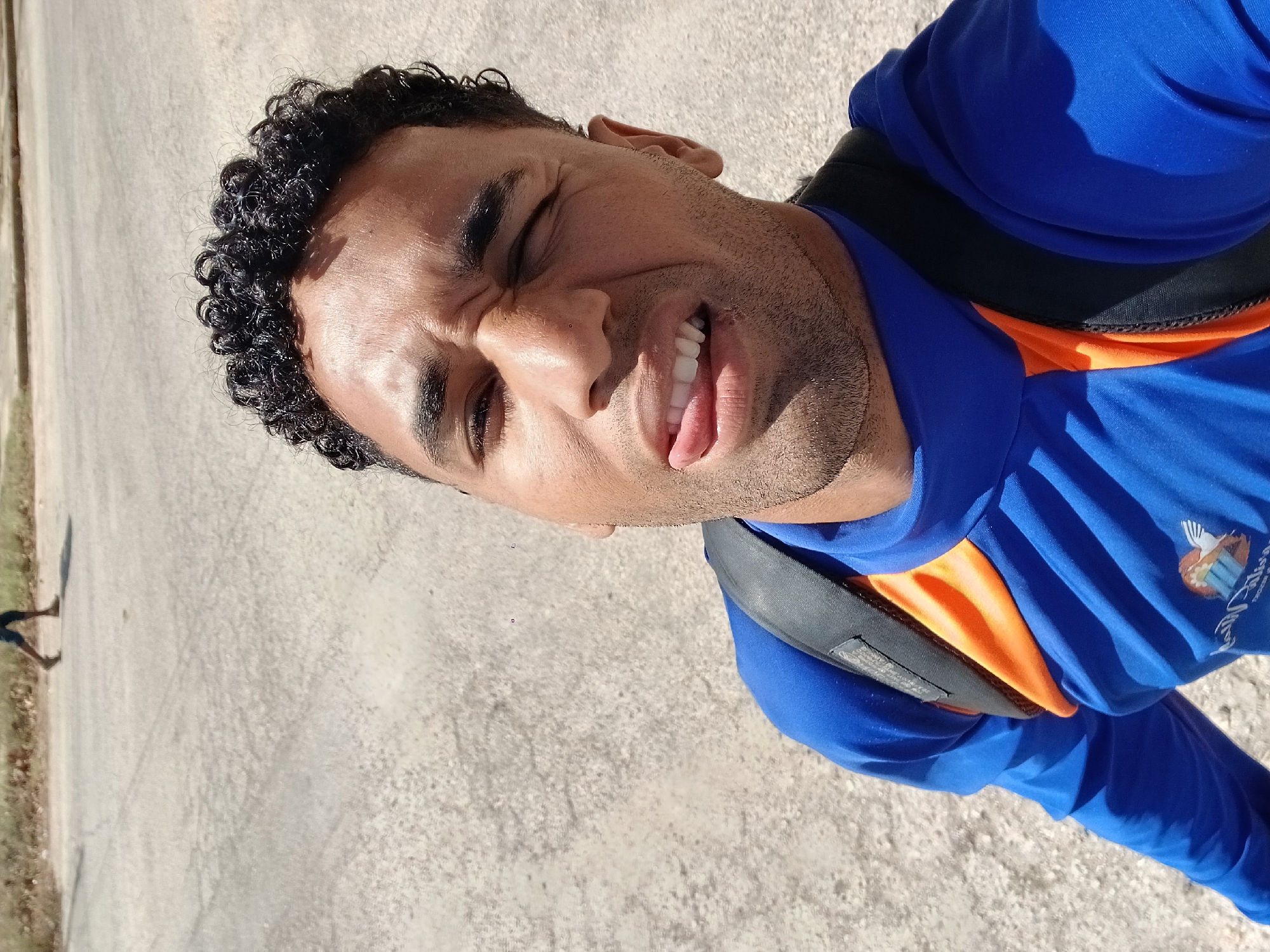 A man is wearing a blue and orange long-sleeved shirt with a black backpack, standing on a paved surface. He has curly black hair and is squinting with his tongue slightly out. The picture appears to be a selfie, with the camera angle looking up at the man. In the lower left corner, there is a blurry figure of a person walking near some vegetation and the edge of the paved area. Without additional information or context, I cannot confirm the location is Porlamar, Venezuela.