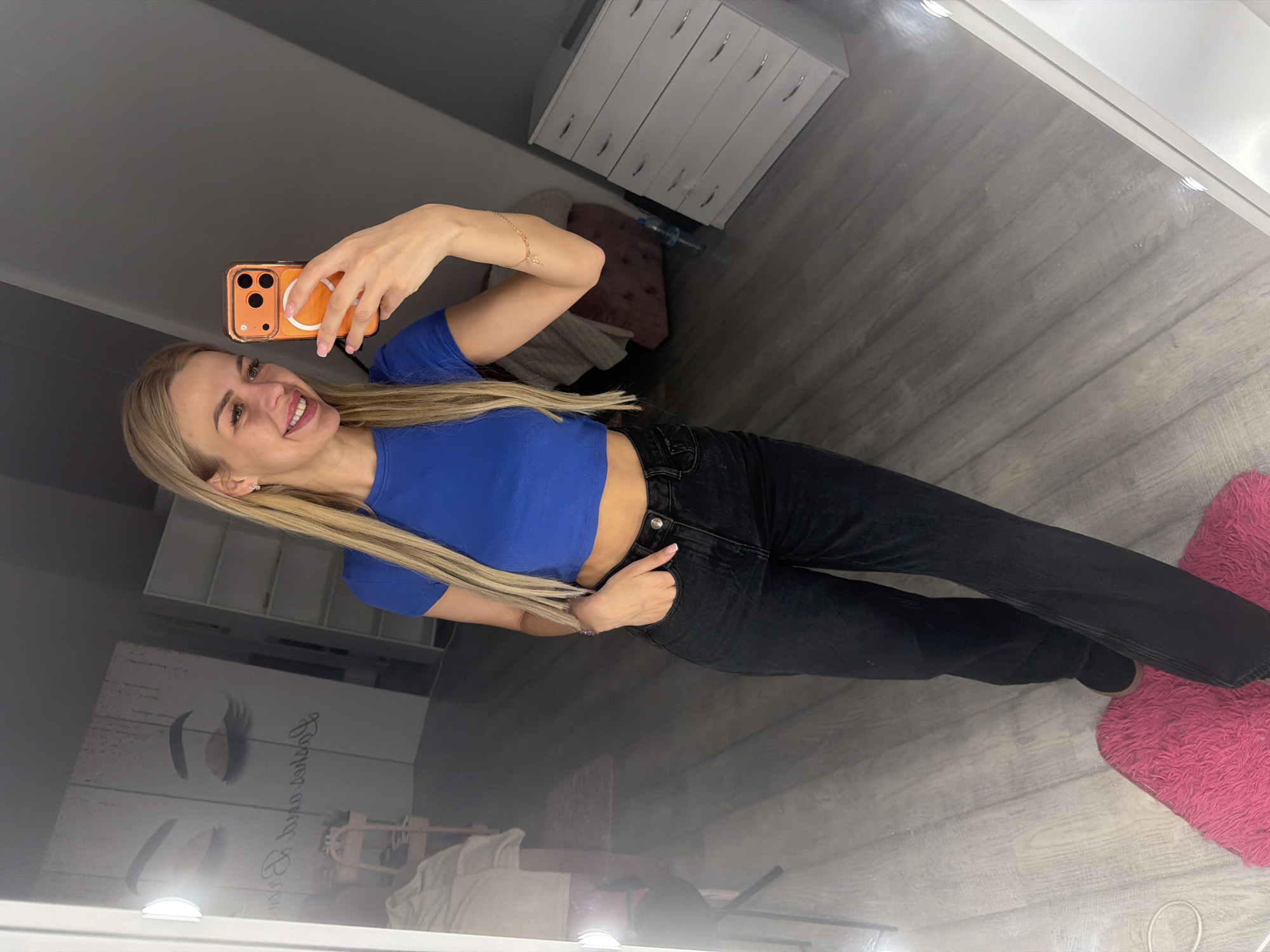 A woman with blonde hair, wearing a blue crop top and black jeans, stands in front of a mirror, taking a selfie with her orange phone. She is smiling and has her left hand in her pocket. Behind her, a white dresser with multiple drawers is visible. To the left, a wall mural depicts closed eyes with eyelashes. The room has a wooden floor. The scene appears to be an indoor space, possibly a salon or dressing room, located in Kyiv, Ukraine.