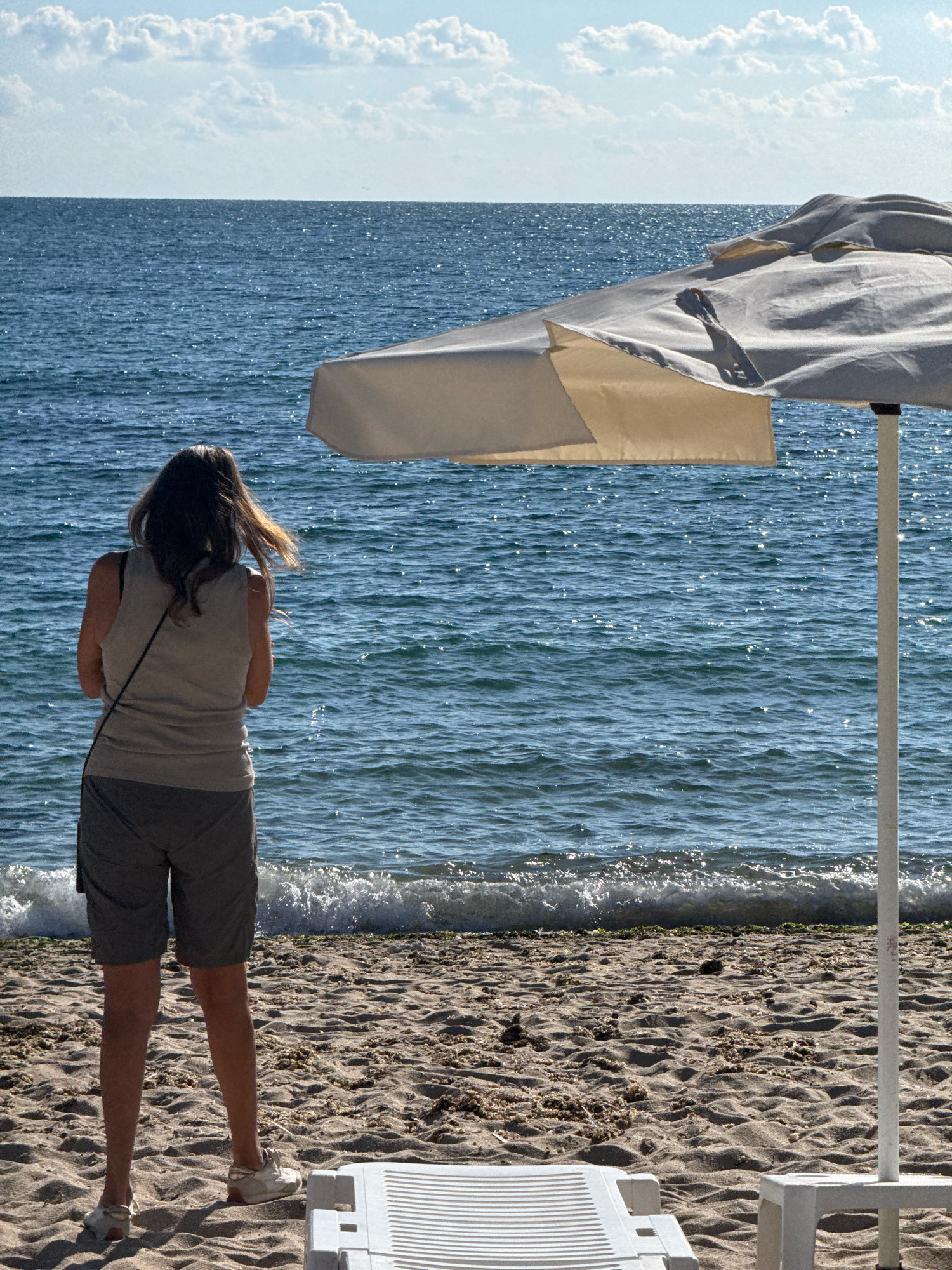 The image depicts a beach scene in Varna, Bulgaria, on a sunny day. In the foreground, a person stands facing the sea, wearing a light-colored sleeveless top and gray shorts. They have dark hair and are wearing white sneakers. To the right of the frame, a large, white beach umbrella is partially visible, casting a shadow on the sand. In the background, the blue sea stretches to the horizon, with gentle waves lapping at the shore. The sky is clear with scattered white clouds. The sandy beach is textured with ripples and some scattered seaweed. A white plastic lounge chair is visible in the lower right corner of the image.