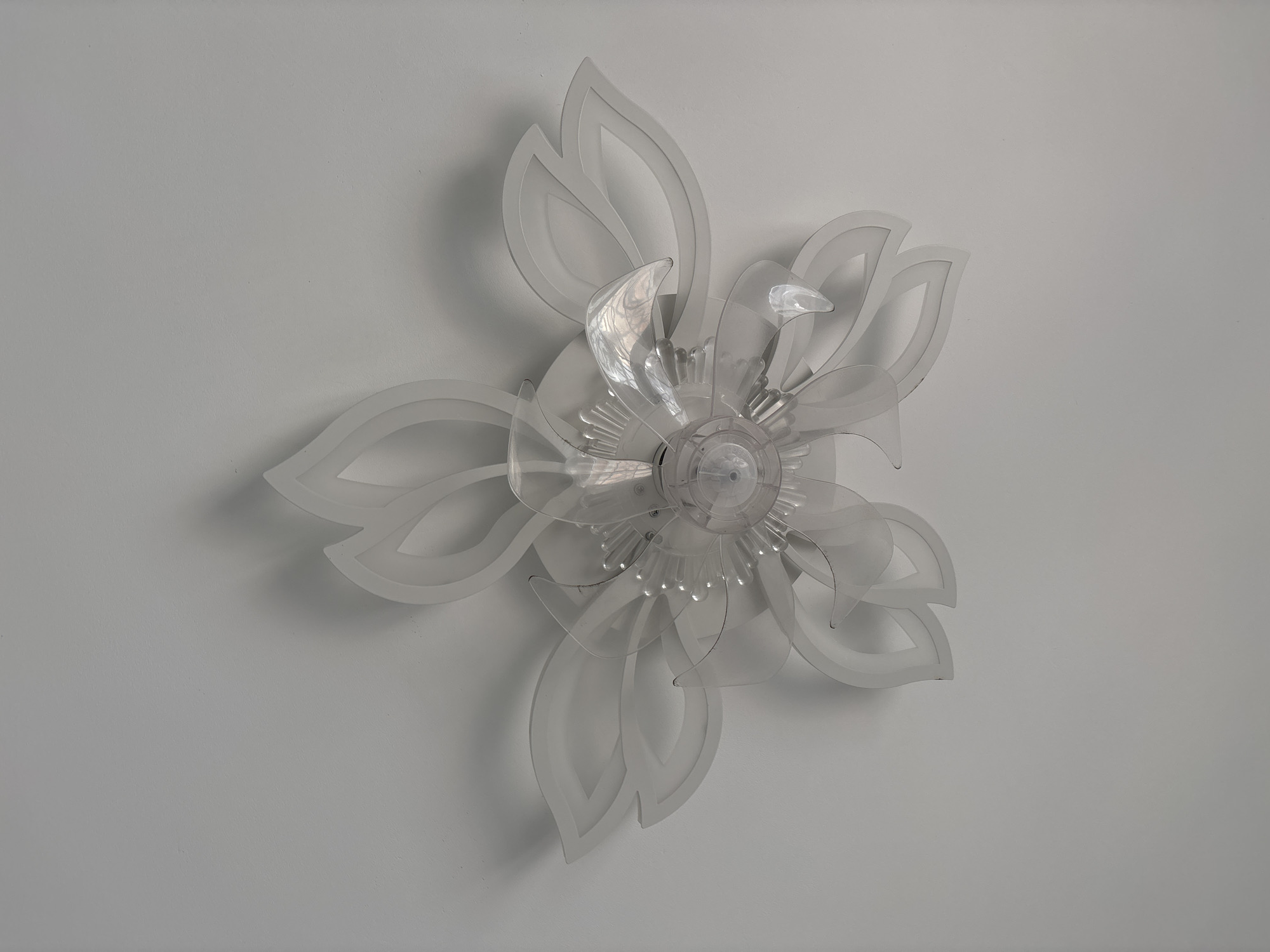 A flower-shaped ceiling fan is mounted on a white ceiling. The fan has multiple translucent white and clear acrylic petals arranged in layers, resembling a blossom. The center of the fan is a clear plastic housing containing the motor and visible screws. The lighting casts soft shadows on the ceiling. This image was taken in Orihuela, Spain.