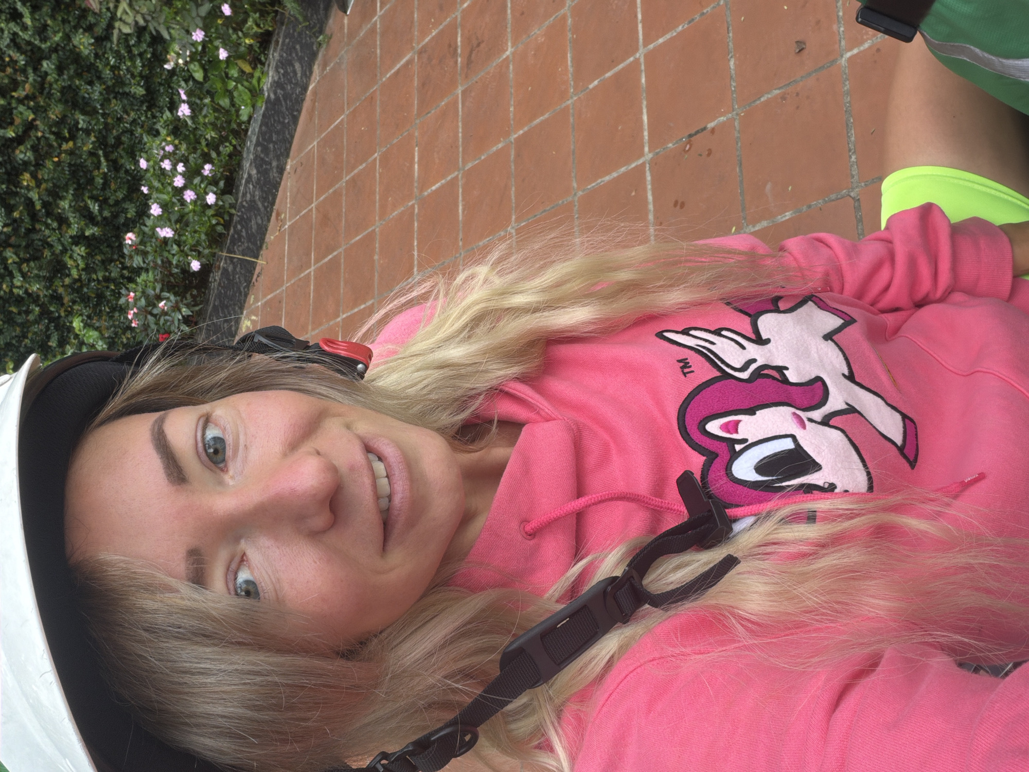 A woman wearing a white helmet and a bright pink hoodie with a cartoon character on it is captured in a selfie. Her blonde hair is visible around her face. She is outdoors, with a paved pathway made of orange tiles in the foreground, and lush green foliage with pink flowers in the background. The scene appears to be in a garden or park setting.