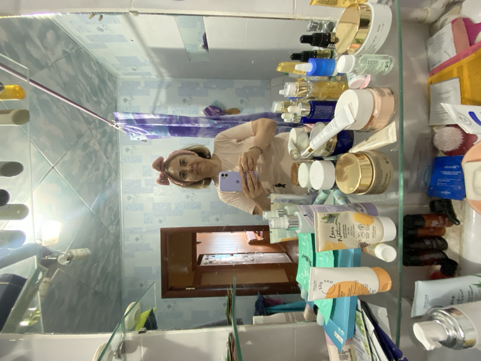 A woman with a pink headband and blonde hair takes a selfie in a bathroom mirror. She is wearing a light pink t-shirt and holding a purple smartphone. The bathroom is tiled with a blue and white pattern on the walls and ceiling. A shower curtain with a purple and blue pattern is visible in the background. The mirror reflects a shelf filled with various beauty products, including bottles, jars, and tubes. In the reflection of the mirror, a doorway leads to another room, revealing a wooden door and a glimpse of furniture. The scene is a personal bathroom, possibly in a residential setting. The location of Urazovo, Russia is not discernible from the visual evidence presented in the image.