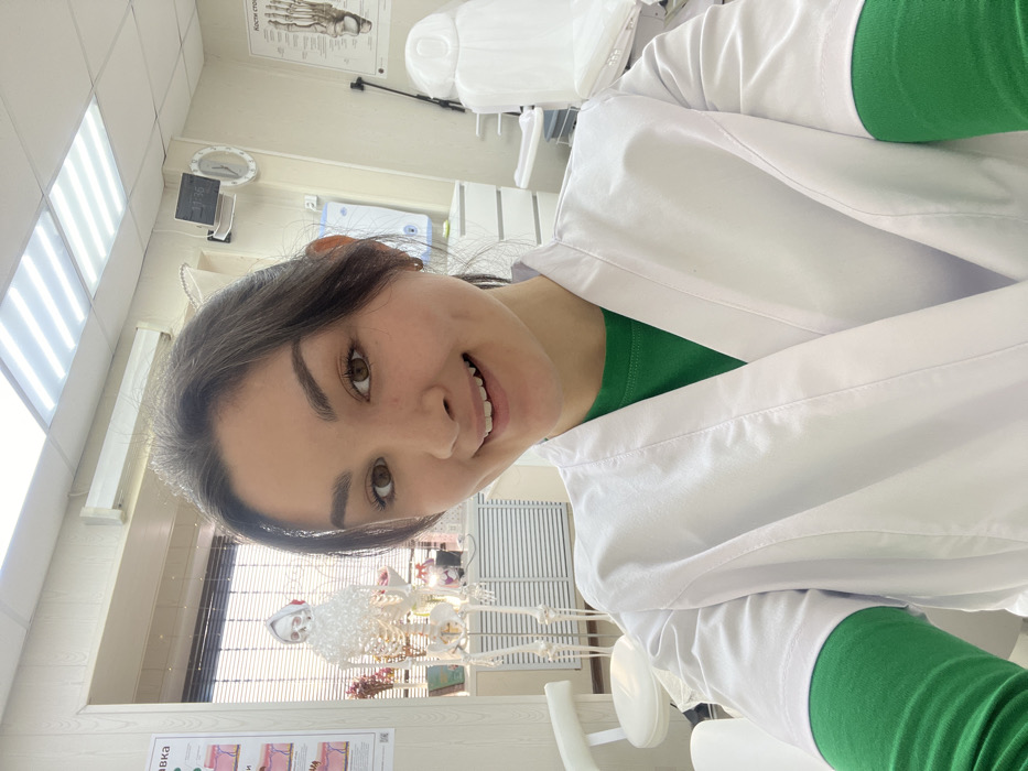 A smiling woman in a white lab coat over a green shirt poses for a selfie in what appears to be a dental office. Behind her are dental equipment including a chair, a skeleton, and posters with anatomical diagrams. The lighting is bright, reflecting off the white walls and equipment in the room. A clock on the wall displays the time.
