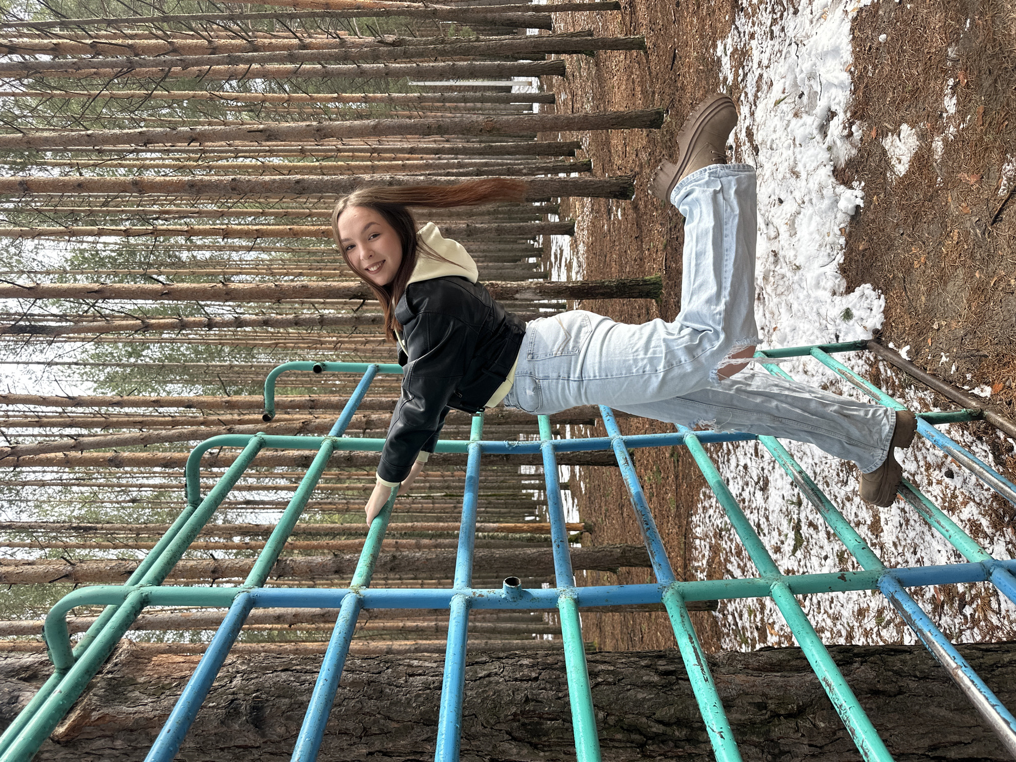 The image shows a young woman lying on a blue and green metal structure, likely a climbing frame or jungle gym, in an outdoor setting in Chaikovsky, Russia. She is wearing a black leather jacket, a white hoodie, light blue jeans, and brown boots. Her hair is brown and extends horizontally across the frame. The background includes bare trees and patches of snow on the ground. The photograph appears to be taken with a slight rotation.