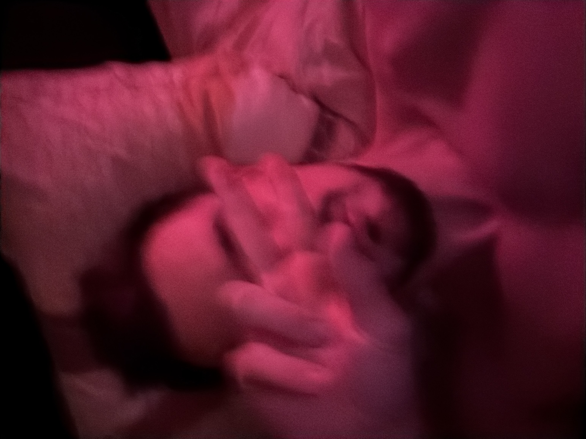 The image is a close-up, blurry shot of a person lying down, possibly in a bed. The lighting is predominantly pink, casting a strong hue over the scene. The person's face is partially obscured by their hand, which is held up in front of their eyes. The background appears to be a pillow or soft surface. There are no discernible elements that would indicate a specific location within the city of Krasnoyarsk, Russia.