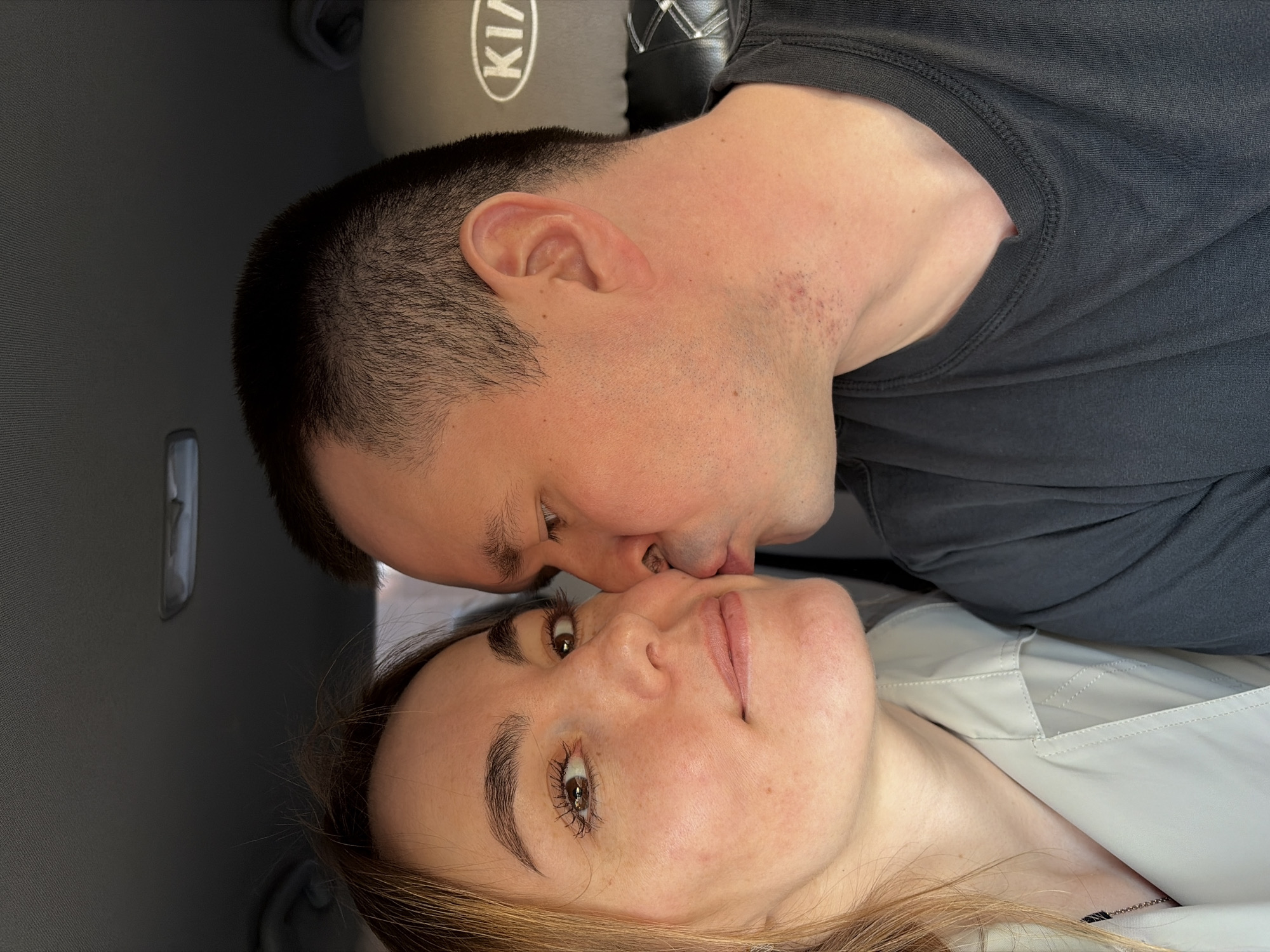 A man and a woman are kissing. The man has dark hair and is wearing a dark gray shirt. The woman has light brown hair and is wearing a white top. They are in what appears to be the back seat of a car, as suggested by the headrest with "KIA" visible. The location in Izhevsk, Russia cannot be verified from the image.