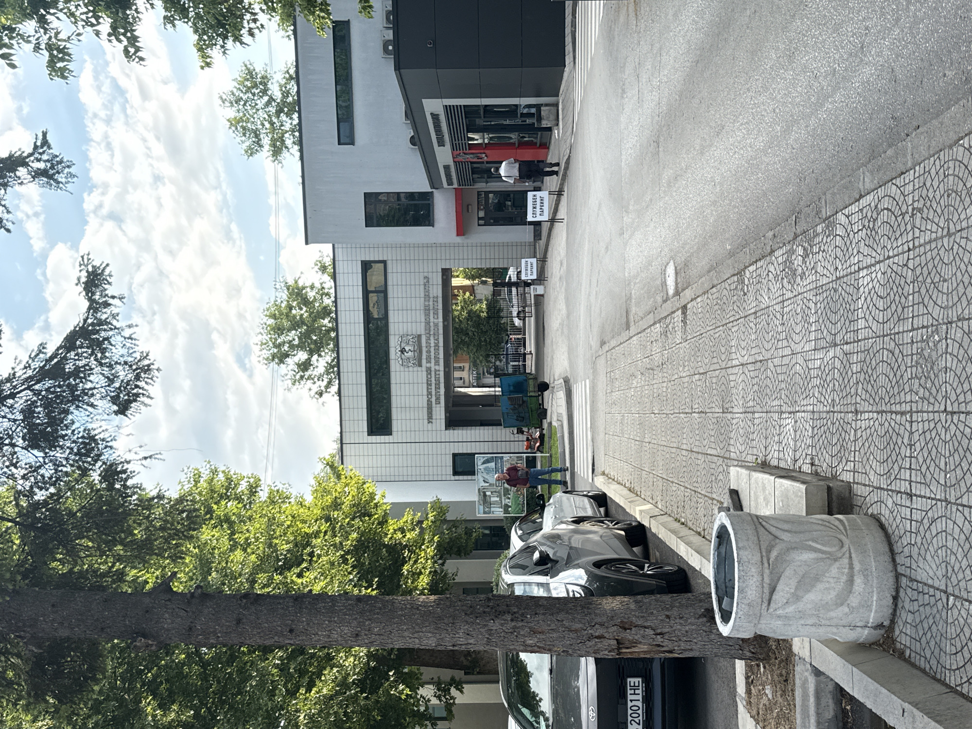 The image shows an exterior view of a building, identified as the University of Food Technologies (UFT) in Plovdiv, Bulgaria. In the foreground, a gray car with license plate "2001 HE" is parked on the left side of a paved road. A large tree trunk is visible in the left portion of the frame. To the right of the car, a sidewalk with a circular patterned tile design runs alongside the road. A concrete planter with a carved leaf motif sits on the sidewalk near the tree.
The building itself is modern, with a white facade and large windows. A sign above an entrance reads "УНИВЕРСИТЕТ ПО ХРАНИТЕЛНИ ТЕХНОЛОГИИ" and "UNIVERSITY OF FOOD TECHNOLOGIES INFORMATION CENTER". The entrance is an archway leading into the building. To the right of this main entrance, there is a smaller, darker-colored building extension with windows and an entrance. Two white signs with black text are placed on the pavement in front of this extension, one reading "СЛУЖЕБЕН ПАРКИНГ" (Service Parking).
Two individuals are visible. One person, wearing a dark jacket and jeans, stands near the main entrance of the building, holding a dark object. Another person, wearing a white shirt and dark pants, is walking away from the camera towards the entrance of the dark-colored building extension. In the background, a blue cart with wheels is visible near a red garden tractor. The sky is partially cloudy. The environment appears to be an urban setting with trees lining the sides of the road and the building complex.