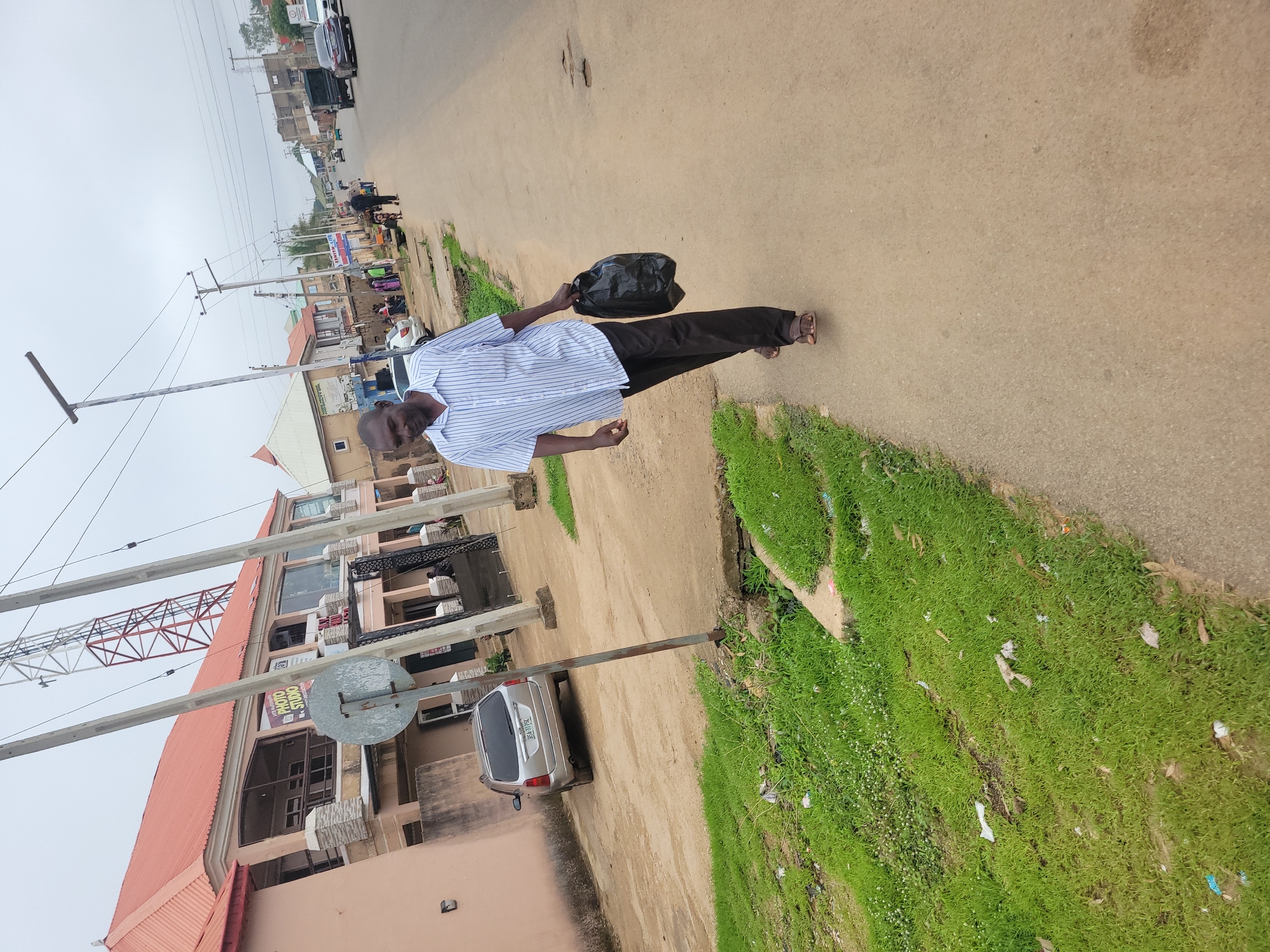 A medium shot captures a man walking along a paved road in Jos, Nigeria. He is wearing a blue and white striped short-sleeved shirt and dark pants, carrying a black plastic bag in his right hand. The man is walking towards the right side of the frame on a wide, somewhat worn asphalt road. To his left, a narrow strip of green grass borders the road, with scattered white and brown debris. Beyond the grass, a sandy path leads to a pale pink building with a red roof. This building features an upper level with a balcony and a sign that reads "PHOTO STUDIO." Further down the road, several other buildings, including a tall, red and white communications tower, are visible under an overcast sky. A silver station wagon is parked on the sandy path near the photo studio building. Other people are seen in the distance along the road and on the sidewalk. The overall environment suggests an urban setting in Jos with ongoing activity.