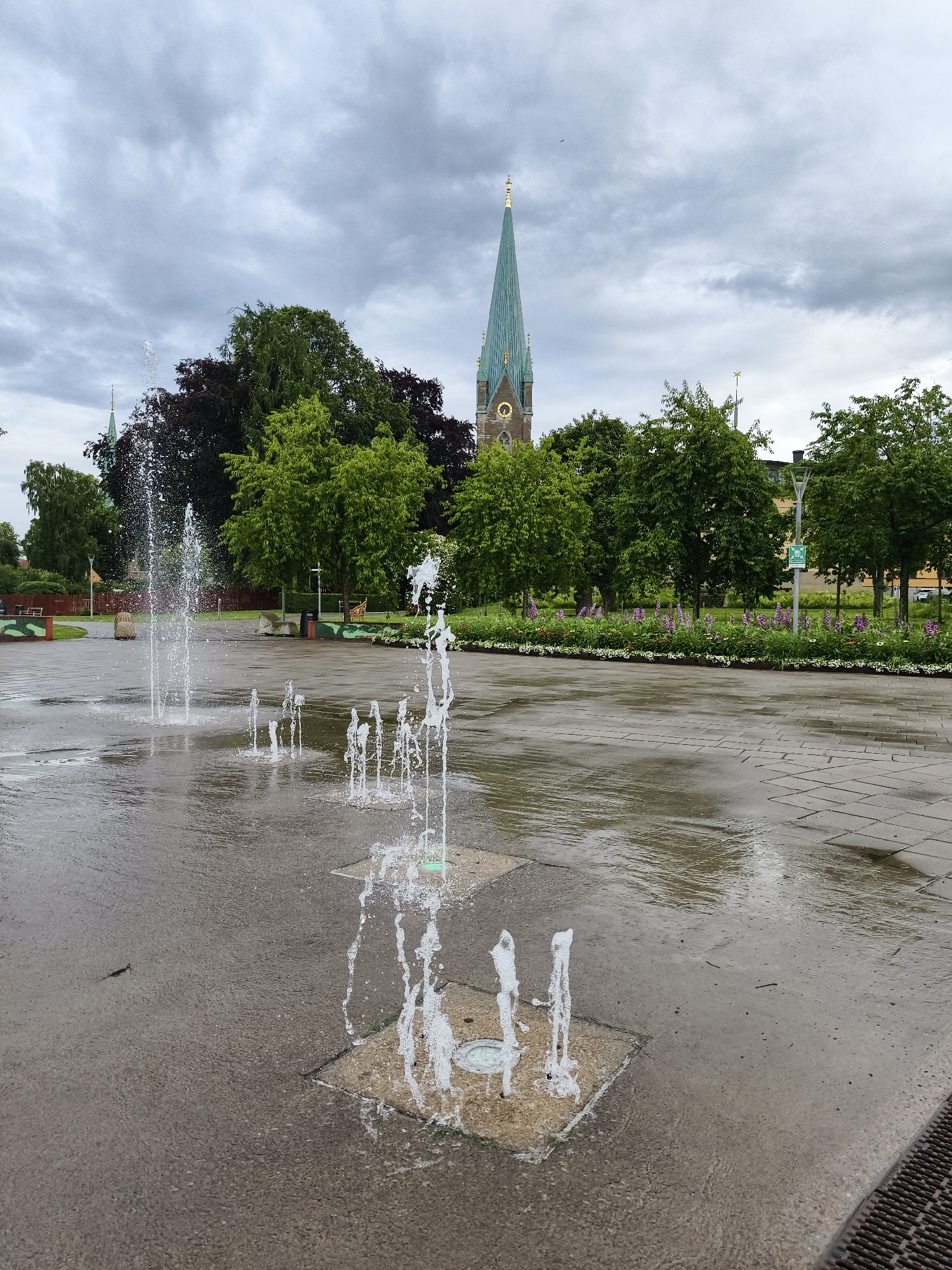 The image depicts a public plaza with an interactive water fountain in the foreground. Several jets of water spray upwards from square recesses in the wet pavement. In the mid-ground, a line of trees separates the plaza from a flowerbed with purple and white blooms. Beyond the trees, a church spire with a gold finial and a clock face is visible against a cloudy sky. The setting appears to be the Stora Torget (Great Square) in Linköping, Sweden, a common public space featuring fountains and adjacent to the Linköping Cathedral. The overall atmosphere is overcast and damp, suggesting recent or ongoing rain. There are no people visible in the frame.
