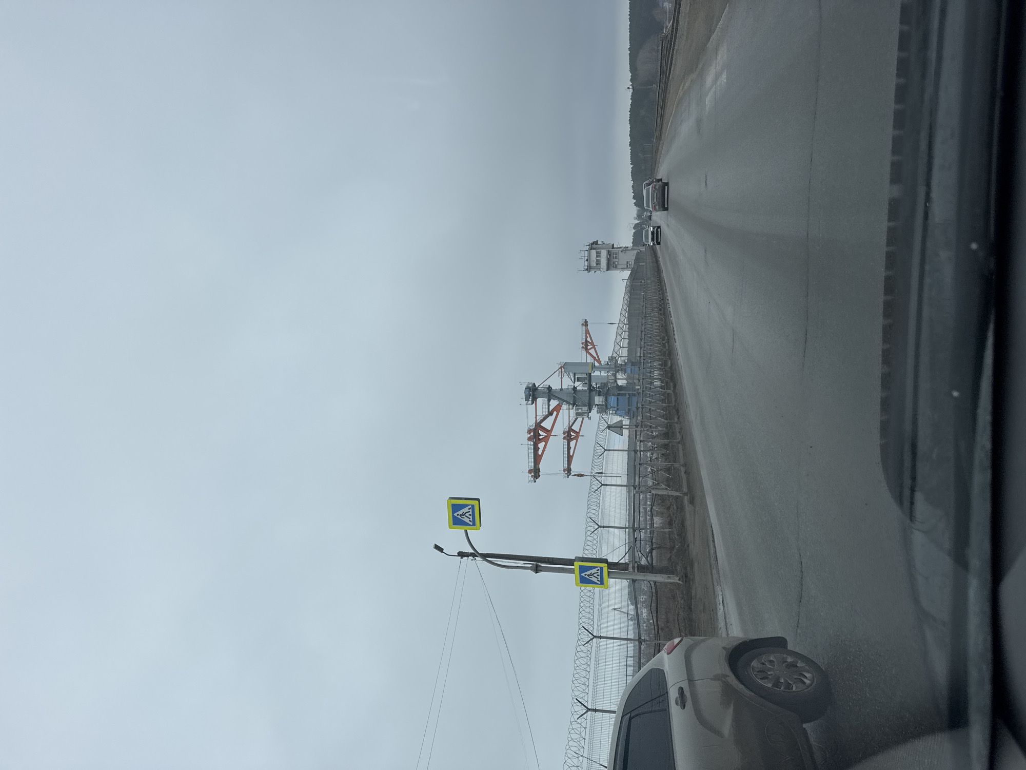 The image shows a road near the industrial area in Chaikovsky, Russia, on an overcast day. The view is captured from inside a vehicle. On the right side of the road, several vehicles are traveling in the same direction. Beside the road is a high security fence with barbed wire. Beyond the fence, there are industrial structures, including cranes. A pedestrian crossing sign is visible near the fence, and a portion of a car is visible in the foreground of the image.