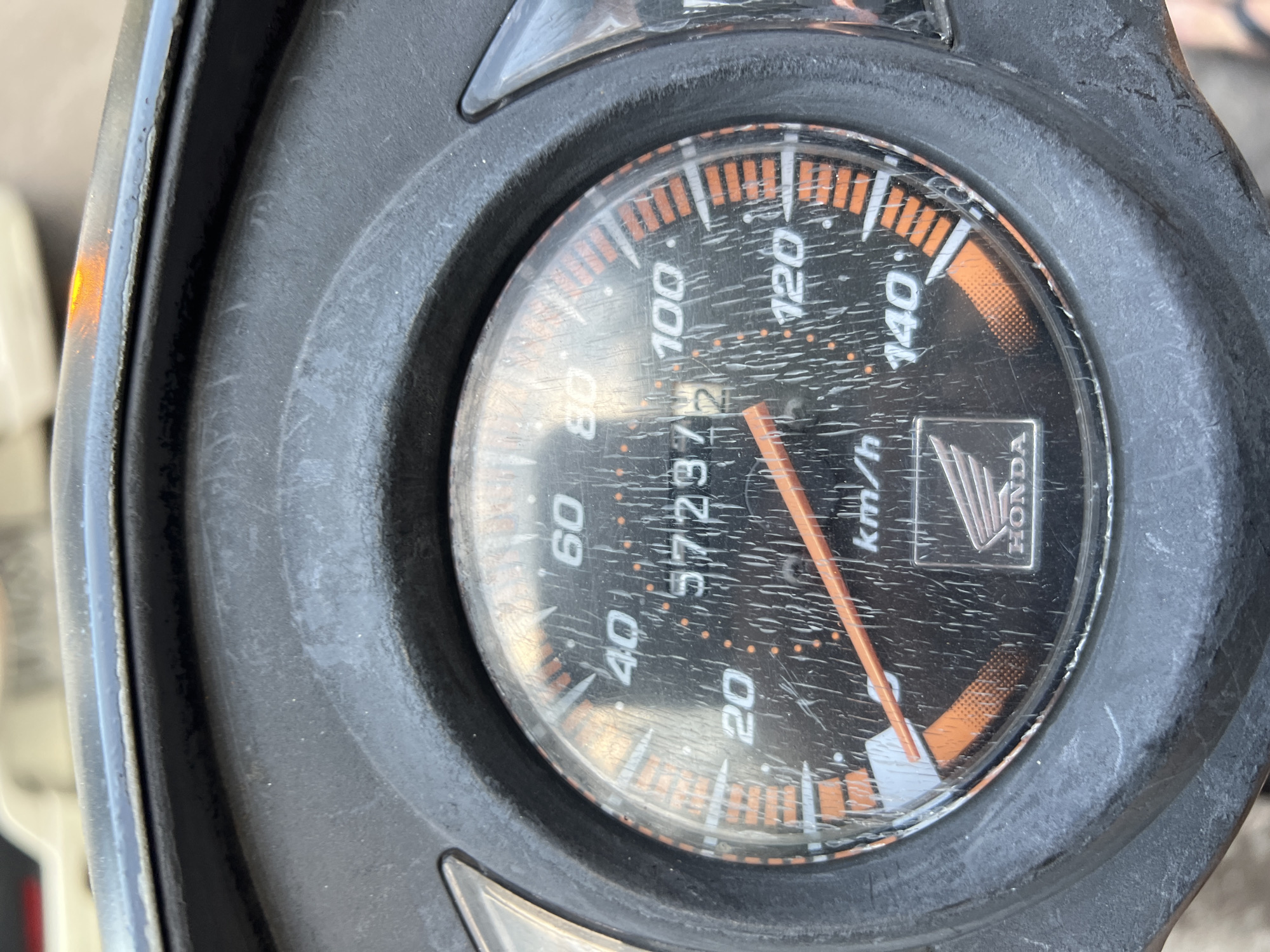 The image is a close-up, eye-level shot of a motorcycle's speedometer. The speedometer is black with orange and white markings. The needle is pointing slightly past 100 km/h. The odometer displays "57237". The Honda logo is visible at the bottom center of the speedometer. The speedometer face is heavily scratched. The surrounding area is out of focus, but appears to be part of a motorcycle dashboard. There are no people visible in the image. The image was taken outdoors during the day, with natural light. The location is Abang, Indonesia, as indicated by the file name.