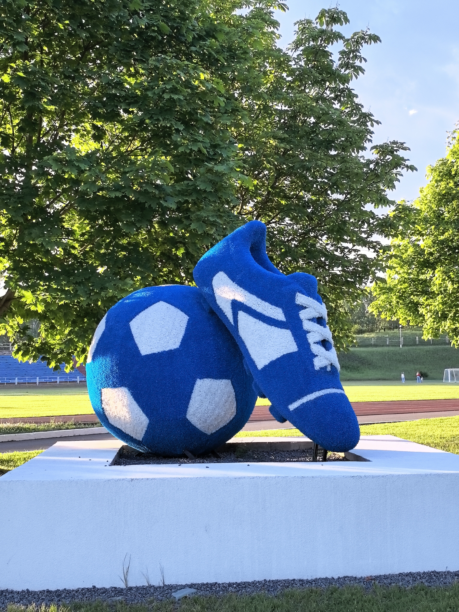 A sculpture of a blue soccer ball and a blue cleat stands on a white pedestal in front of a stadium. The soccer ball is large and round, with white pentagons scattered across its surface. The cleat is leaning against the ball, with white accents forming stripes and laces. Both the ball and the cleat are covered in a fuzzy, plush material. Behind the sculpture, stadium bleachers are visible, as well as a track and a grassy field. Several trees with green leaves frame the top and sides of the image. The sky is blue with a few scattered clouds. The scene is set outdoors in what appears to be a sports complex. There are a few distant, indistinct figures visible on the track in the background.