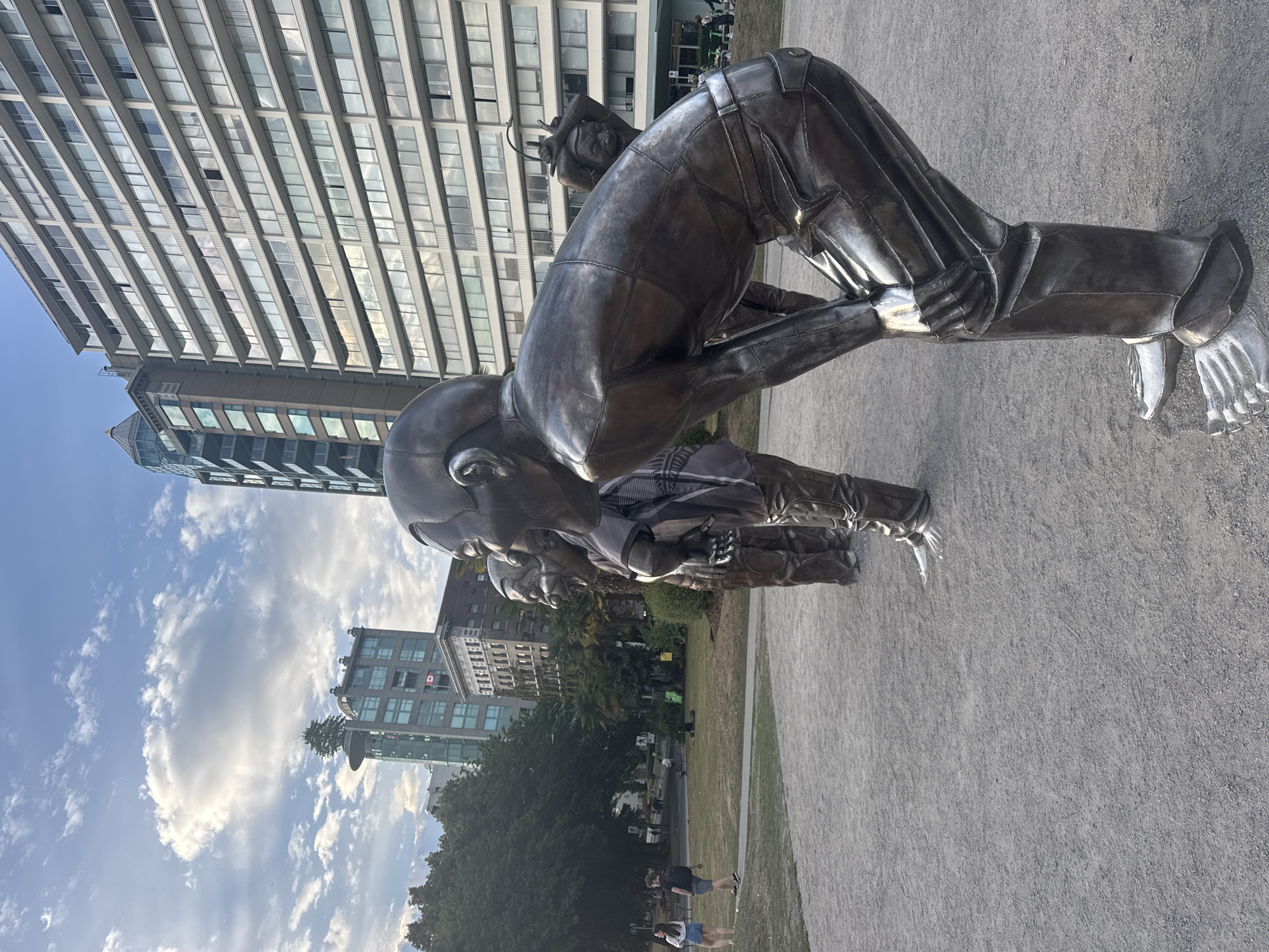 In a park in Vancouver, Canada, at least four large bronze sculptures of figures are positioned in a row, all leaning forward with their heads down, as if looking at something on the ground. The figures appear to be in motion, with exaggerated facial expressions of laughter. They are metallic, with visible seams from their construction, and wear clothing that resembles pants and shirts. The foreground is a gravel path, and the background features a large, modern building with many windows, as well as trees and other parkgoers. The sky is partly cloudy, with a mix of blue sky and white clouds. The light suggests it is either late afternoon or early morning.