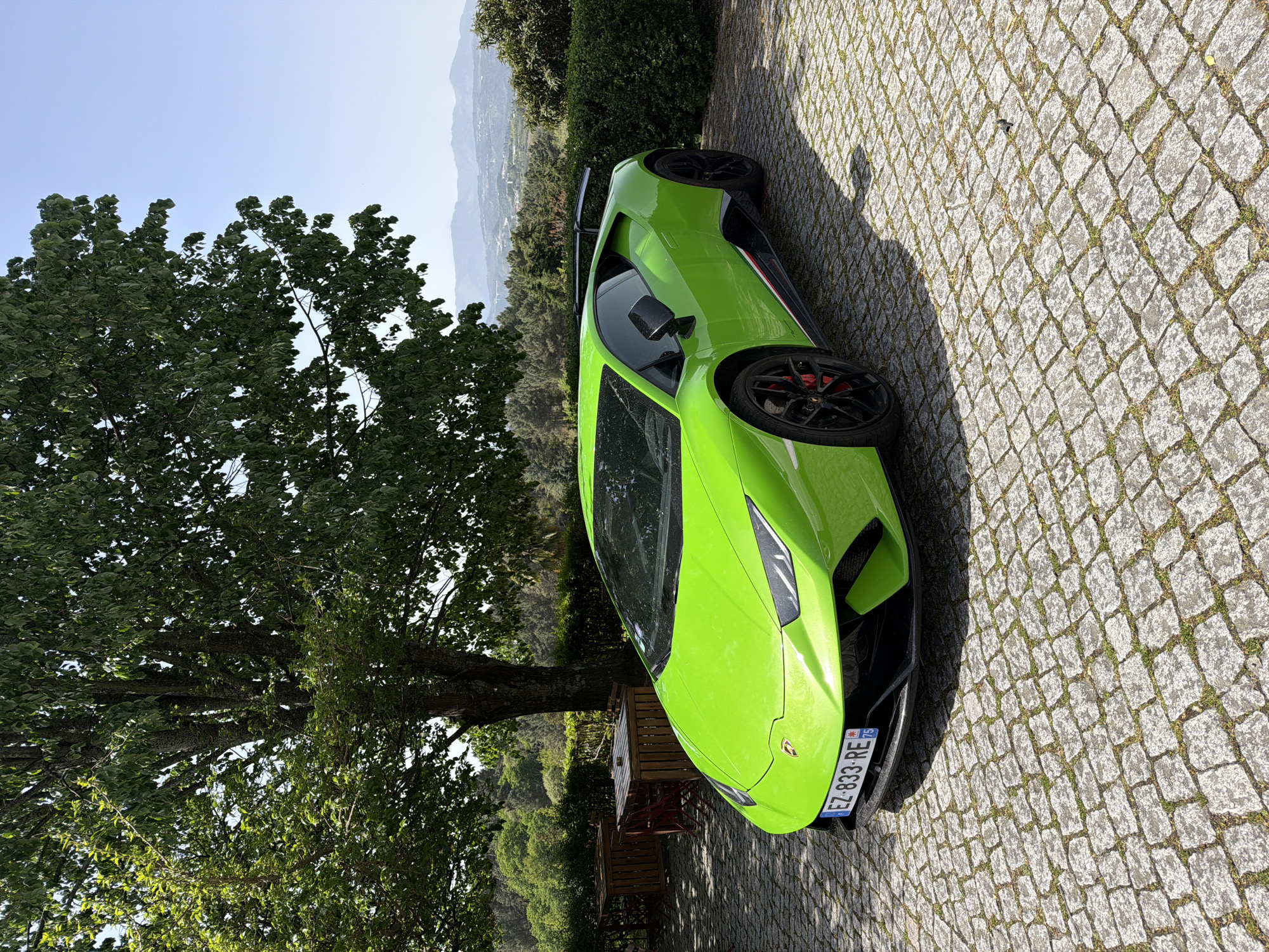 A bright green Lamborghini Huracan is parked on a cobblestone driveway. A large tree stands to the left of the car, providing partial shade. In the background, a green hillside and distant mountains can be seen. The license plate of the car is visible and partially legible. The image does not indicate it is located in Vila Real, Portugal. The location cannot be determined from the image.