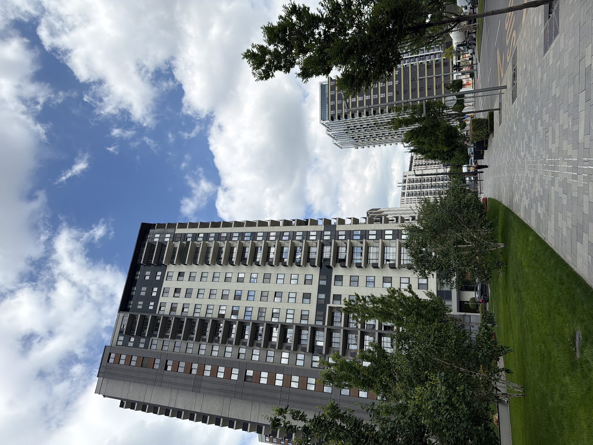 The image shows a cityscape in Belgrade, Serbia, featuring a tall, modern apartment building as the primary subject. The building is multi-story, with a mix of beige and dark gray facade, and incorporates recessed balconies on many floors. A mix of blue sky and white clouds forms the background to the building. In the foreground, there is a patch of green grass, and several trees. To the right, there is a paved sidewalk and a street with sparse traffic. In the distance, there are other tall buildings, continuing the urban landscape. The photo is taken from a low angle, emphasizing the height of the main building.