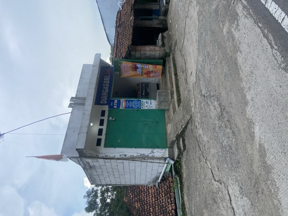 The image shows a small storefront in Cipatat, Indonesia. The store is located at a crossroads, visible at the bottom of the image. It is a one-story concrete building with a green metal door. The storefront has a sign above the door that says "Diargasbrilink" and another sign indicating an ATM. An orange flag is attached to a pole on the roof. To the right of the store is a sign advertising “Es Teh Jumbo Rp. 3000” with a photo of iced tea. The storefront also has a small window with goods visible. The store has a cement step, a ramp, and a cement pad. The road in front of the store is paved and cracked. To the right, a second building is visible with a tiled roof. Vegetation is visible in the background. There are no people visible. The store is in the city of Cipatat, Indonesia. The coordinates are -6.778 latitude and 107.437 longitude.