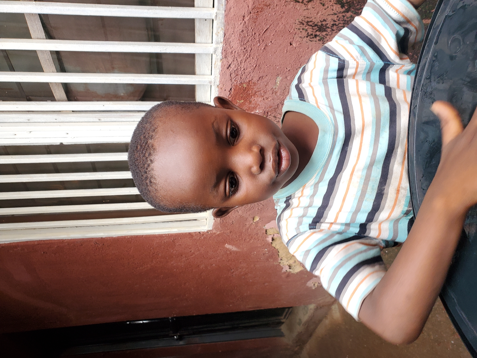 A young Black boy is shown from the chest up, looking down to his left with a slight pout. He has short, dark, curly hair and dark brown eyes. He is wearing a short-sleeved, horizontally striped t-shirt in shades of white, light blue, dark blue, and orange. His left arm is bent and rests on a black object, possibly a bucket or basin, which is in the foreground. The background consists of a textured, reddish-brown wall with peeling paint and a barred window with cream-colored frames. The overall setting appears to be an outdoor area or a poorly maintained building, consistent with some urban environments in Jos, Nigeria.