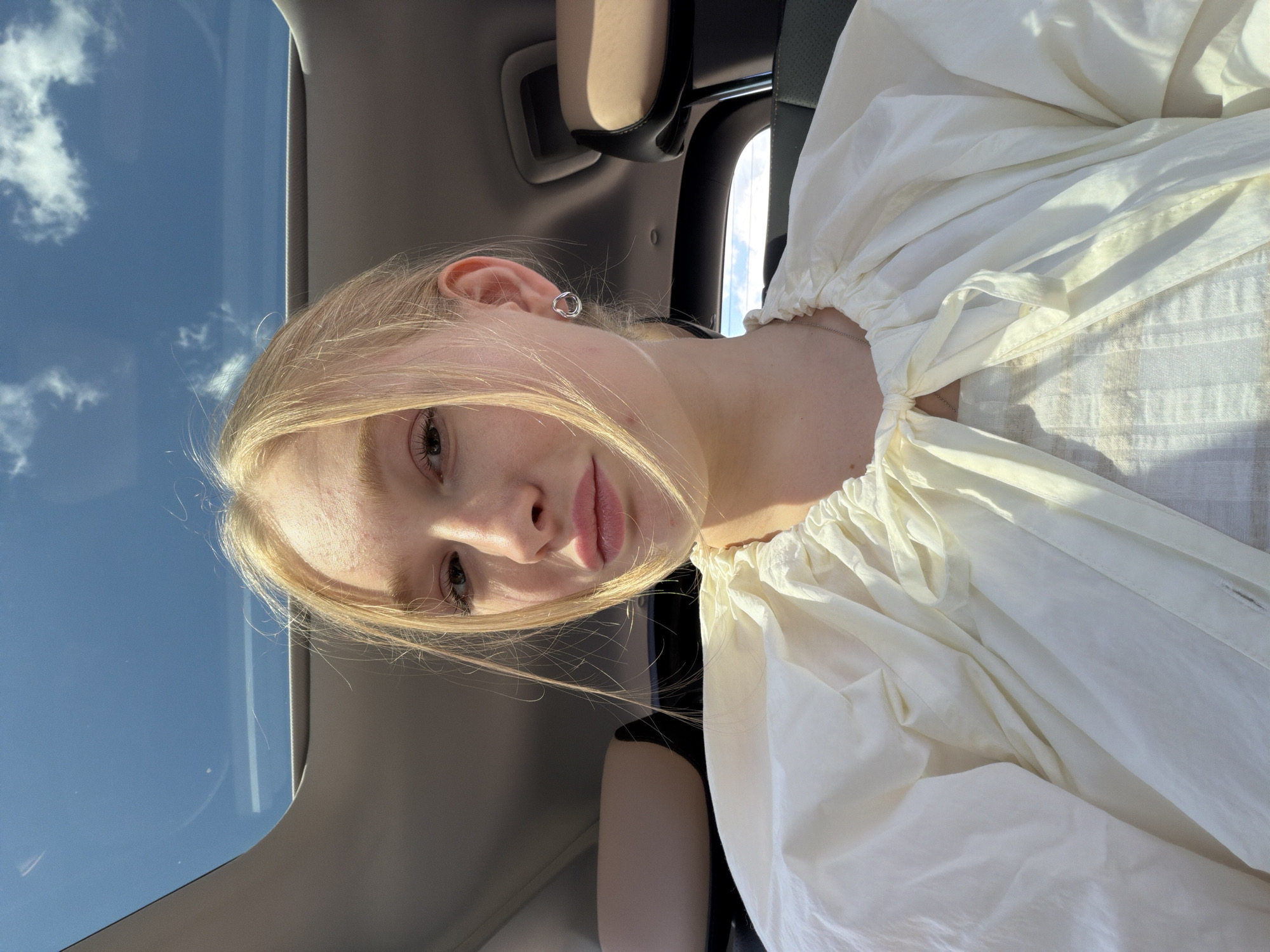 This media file is a portrait of a young woman with blonde hair, taken from a slightly angled, close-up perspective. She is wearing a white, voluminous blouse with gathering at the neckline. Her left ear is visible, adorned with a small, circular silver earring. Her skin is fair and appears illuminated by natural light, casting subtle shadows on her face. Her gaze is directed towards the camera.
The background suggests an indoor setting, likely a car, with hints of car interior elements such as a headrest and a dark seat. A significant portion of the image is dominated by the sky, visible through what appears to be a sunroof or window. The sky is bright blue with scattered white clouds, indicating it is daytime with clear weather.
The overall mood appears casual and relaxed, possibly a selfie taken during a car ride. The lighting suggests it is either morning or afternoon, given the brightness of the sky. No text is visible in the image. The provided location context is Rossiyskiy, Russia.