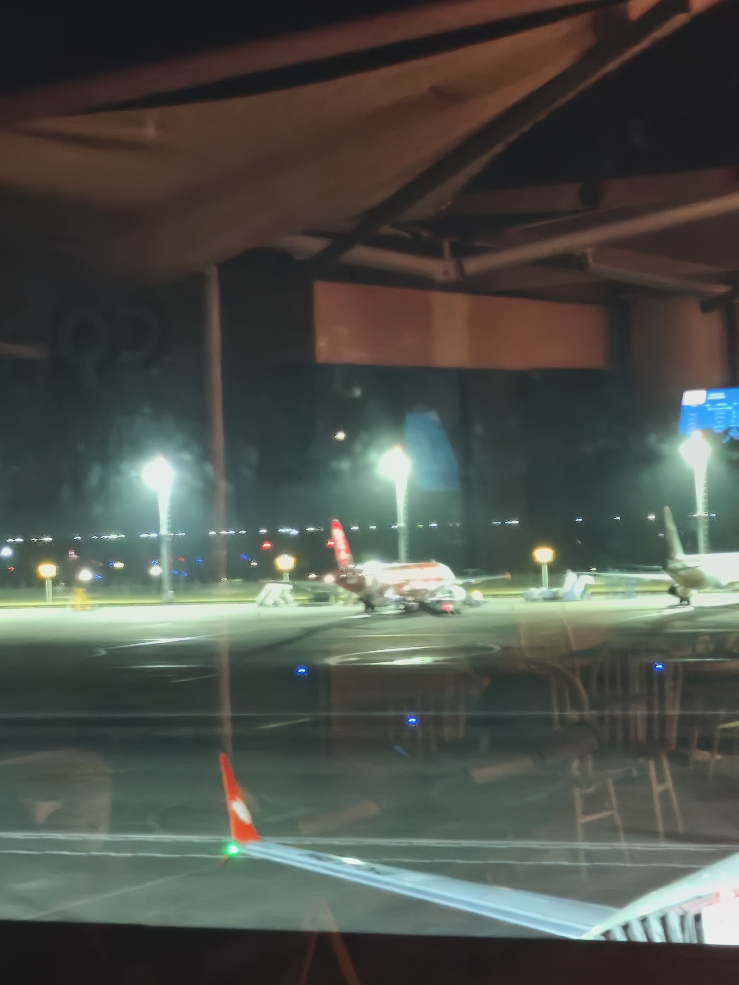 The image depicts an airport tarmac at night. The scene is dimly lit by runway lights and spotlights, creating a nocturnal atmosphere. Several airplanes are visible on the tarmac. One prominent airplane in the mid-ground has a red tail and white fuselage. Another airplane, partially visible on the right, is light-colored. In the foreground, the wing of an aircraft is visible, featuring a red and white tip with a green light. The airport environment suggests a busy hub, with various lights and structures indicating infrastructure for air travel. The overall impression is one of a functioning airport during nighttime operations. There are no visible people, and no discernible text. The weather appears to be clear, as there are no indicators of rain or fog. The location context of Kuta, Indonesia, places this scene in a tropical region, though this is not directly evident from the image itself.