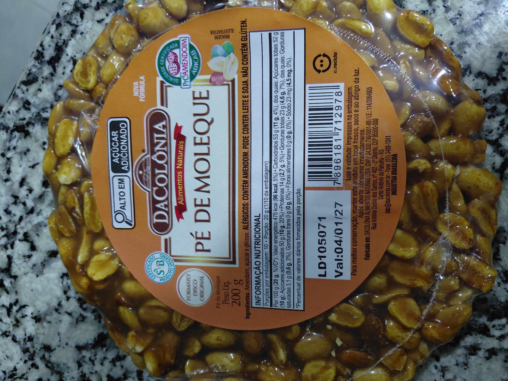 The image shows a close-up of a package of "Pé de Moleque," a Brazilian peanut candy, with a circular orange label. The label features branding for "DACOLONIA Alimentos Naturais." Text on the label indicates it's a vegan product with added sugar, and lists nutritional information. The product is a disc-shaped confection composed of peanuts bound together with a caramel-like substance. The background is a speckled countertop. There is no information within the image that relates to the city of Encarnacion, Paraguay.