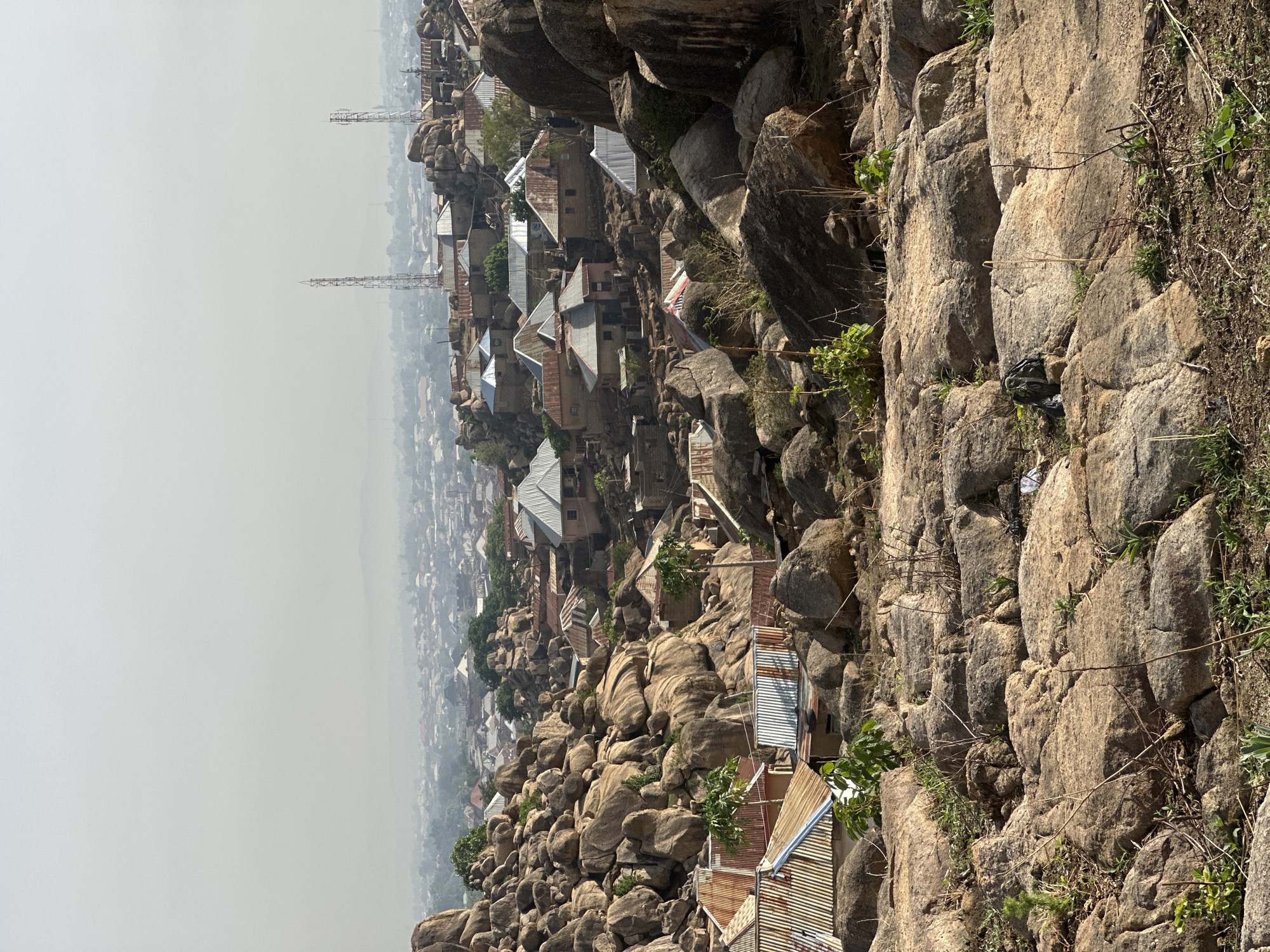 This photograph shows a view of a settlement nestled among large rocks, likely located on or near a rocky outcrop or hill. The location is identified as Jos, Nigeria. The houses appear densely packed, with visible roofs, some made of corrugated metal. There are several communication towers visible in the background. The foreground is dominated by large, irregularly shaped rocks and sparse vegetation. The background shows a hazy cityscape.