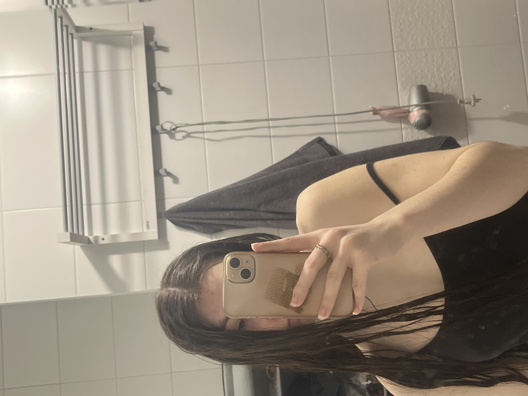 The image is a selfie taken in a bathroom, likely with a mobile phone. The bathroom features white tiled walls, a towel rack, and a hair dryer mounted on the wall. A gray towel is draped nearby. A person is holding the phone, obscuring part of their face. They have long, dark hair. Their skin is fair and they are wearing a black top. The location cannot be determined from the image and there is no indication it is in the city of Saint Petersburg, Russia.