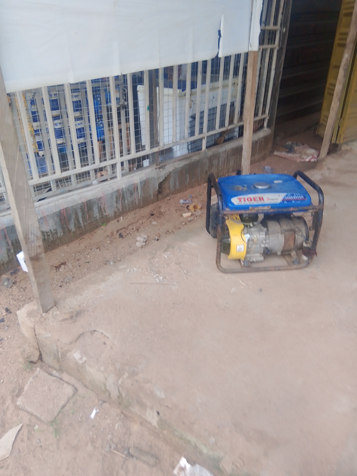 The image depicts a portable generator with a blue top and a yellow fan, situated on a concrete surface in front of a building with a white overhang and a barred window. The ground is a mixture of concrete and dirt, with some debris scattered around. In the background, through the bars, stacks of blue and white items are visible, possibly merchandise or construction materials. The scene appears to be an outdoor setting, possibly a shop or a workshop, in the city of Jalingo, Nigeria, given the context provided. There are no people visible in the image.