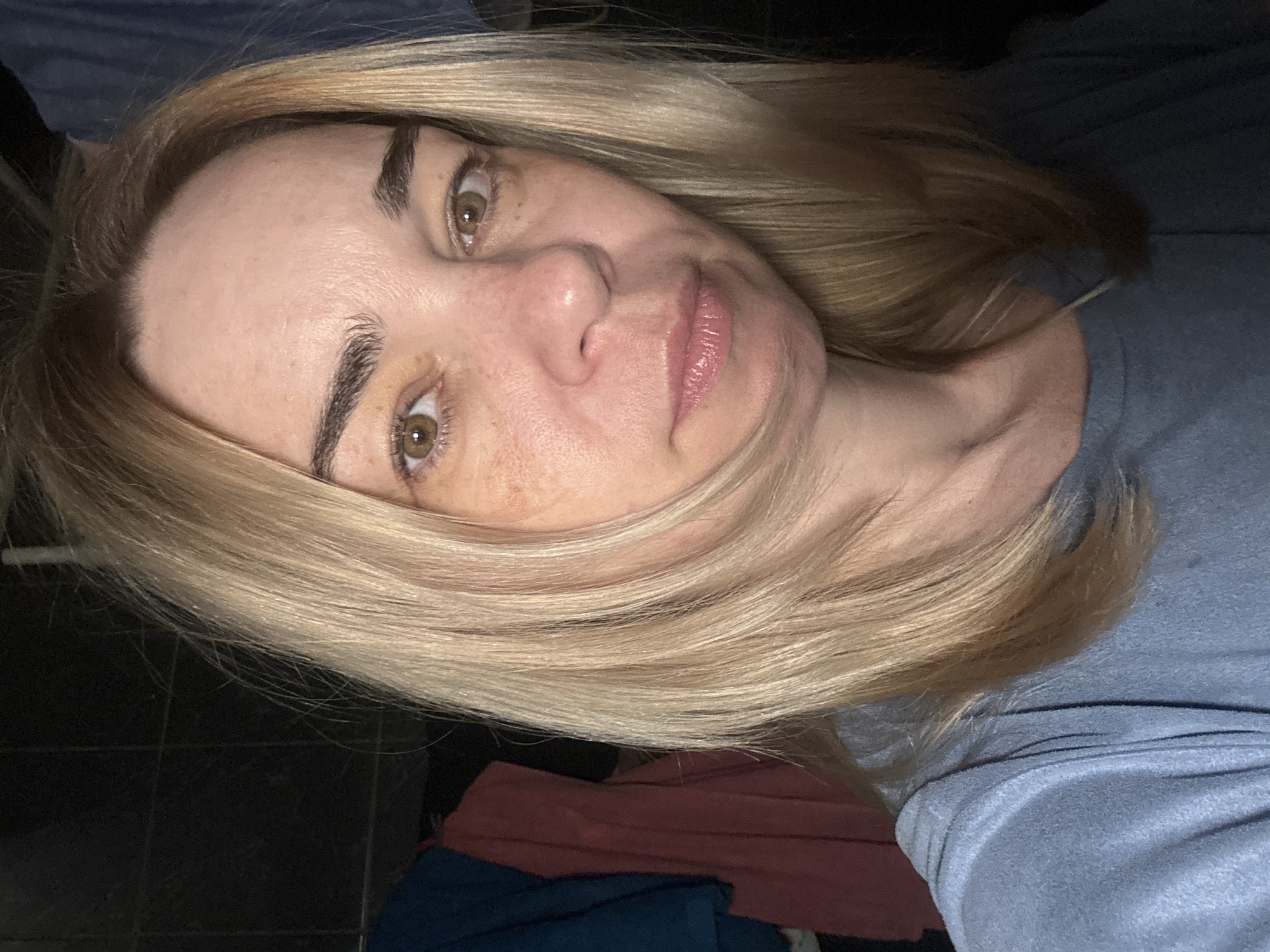 A close-up, slightly angled selfie of a middle-aged Caucasian woman with fair skin, freckles, and hazel eyes. She has light blonde, shoulder-length hair. She's wearing a light blue, long-sleeved shirt. Background shows dark tile flooring and a glimpse of fabric in burgundy and dark blue. No indication of Vsevolozhsk, Russia can be determined.