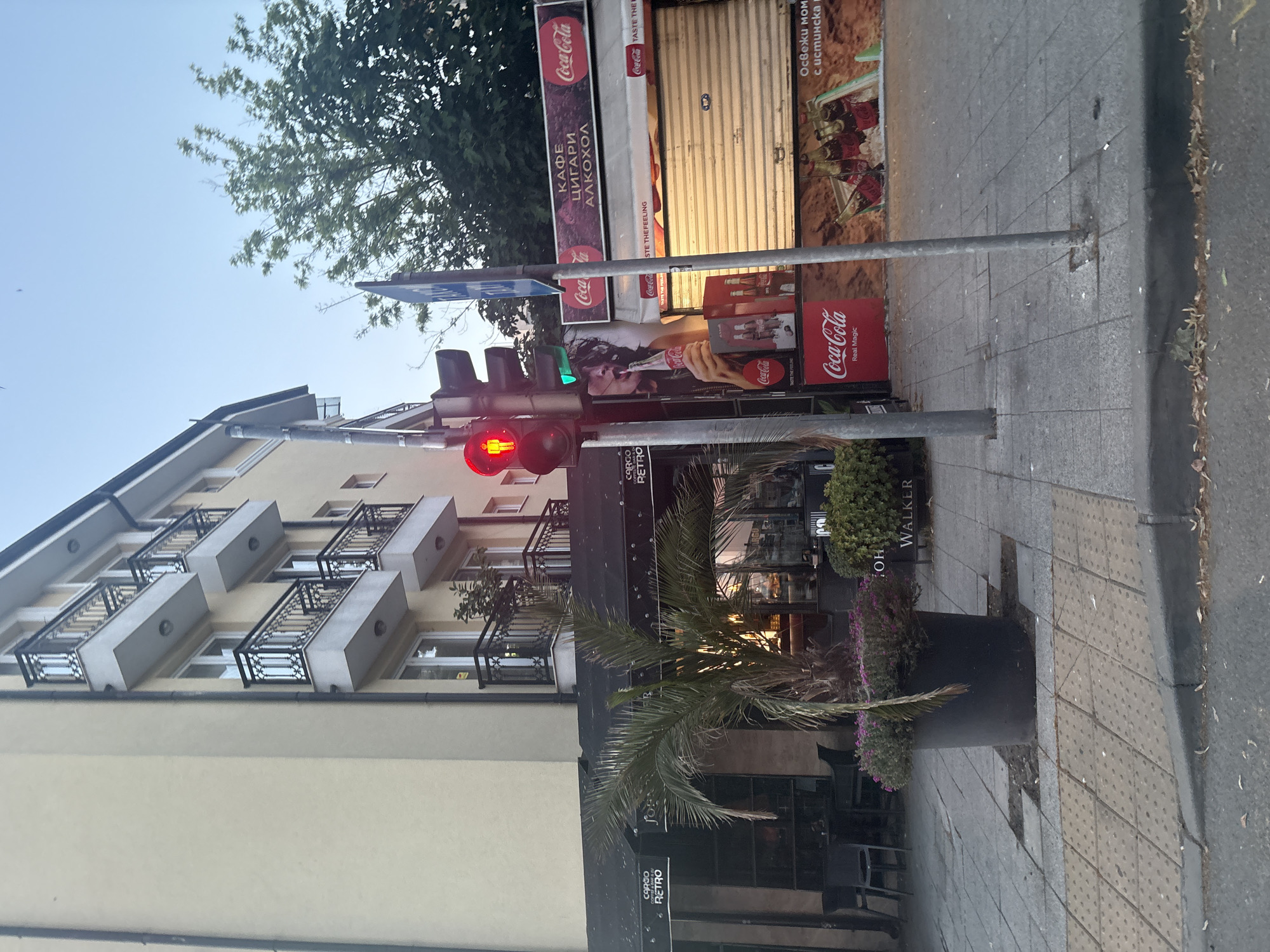 The image depicts a street scene in Burgas, Bulgaria, likely on a sidewalk next to a building with commercial establishments. A traffic light is prominently displayed, showing a red pedestrian signal and a green vehicular signal. Adjacent to the traffic light, a Coca-Cola branded kiosk or shop is visible, featuring advertisements for the beverage and possibly alcohol. A palm tree is planted in a large pot on the sidewalk, and there are outdoor seating areas for a cafe or restaurant. The building appears to be multi-story with balconies. The overall impression is that of an urban commercial area, possibly in the evening or early morning due to the lighting conditions.