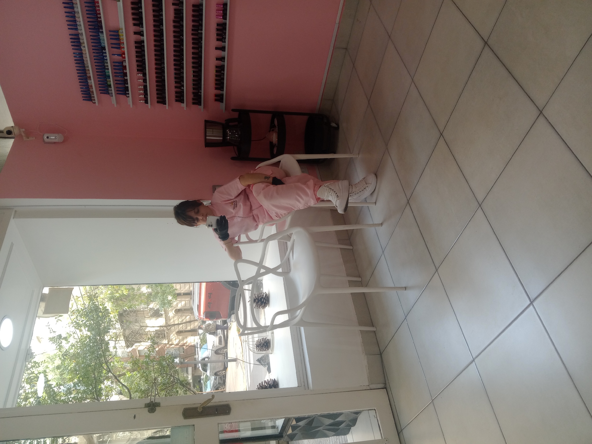 A full shot reveals a woman, presumably a young adult, dressed in a pink suit and white sneakers, sitting on a white chair indoors. She is holding a black object, possibly a cell phone, in her hands. The background features a pink wall adorned with multiple shelves displaying various nail polish colors. Adjacent to the wall, there is a black object on the floor. The scene appears to be a nail salon or beauty parlor. The location appears to be Buenos Aires, Argentina, as the street seen through the window suggests this location. Outside, there is an urban street with buildings, trees, and parked cars. The doorway is open, providing a view to the street.