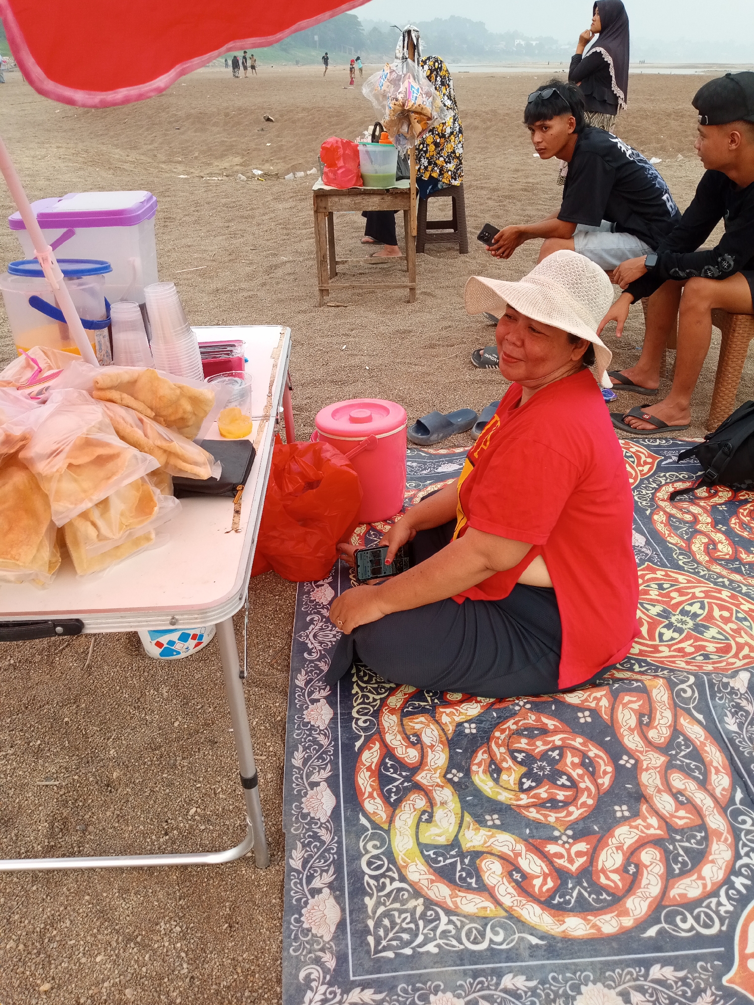 A woman in a red shirt and straw hat sits on a patterned mat on a sandy beach. She is looking to her left, with her hands resting on her lap. In front of her is a small table with various items including plastic containers, cups, and bags of food. Behind her, two young men are seated, one of whom is looking at his phone. Further in the background, other people are visible on the beach, and a body of water can be seen. The scene appears to be a vendor stall at a beach in Kapuas, Indonesia.