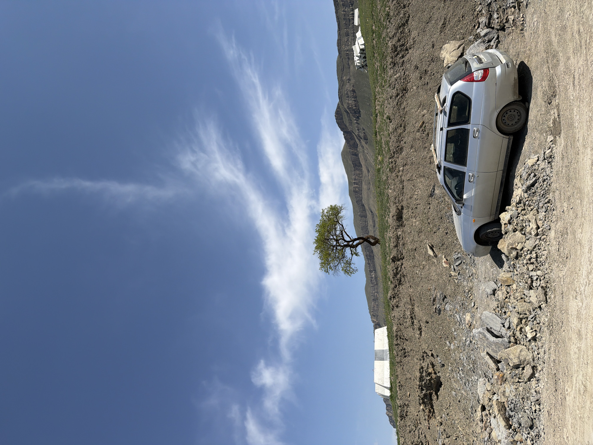 A silver station wagon is parked precariously on the edge of a rocky dirt road in a mountainous area. The ground is barren and covered with loose rocks and sparse vegetation. A lone tree with green leaves stands further up the slope. In the background, a mountain range is visible under a blue sky with wispy clouds. A concrete barrier with a black and white checkerboard pattern is situated near the edge of the road. The city and specific location are unknown.