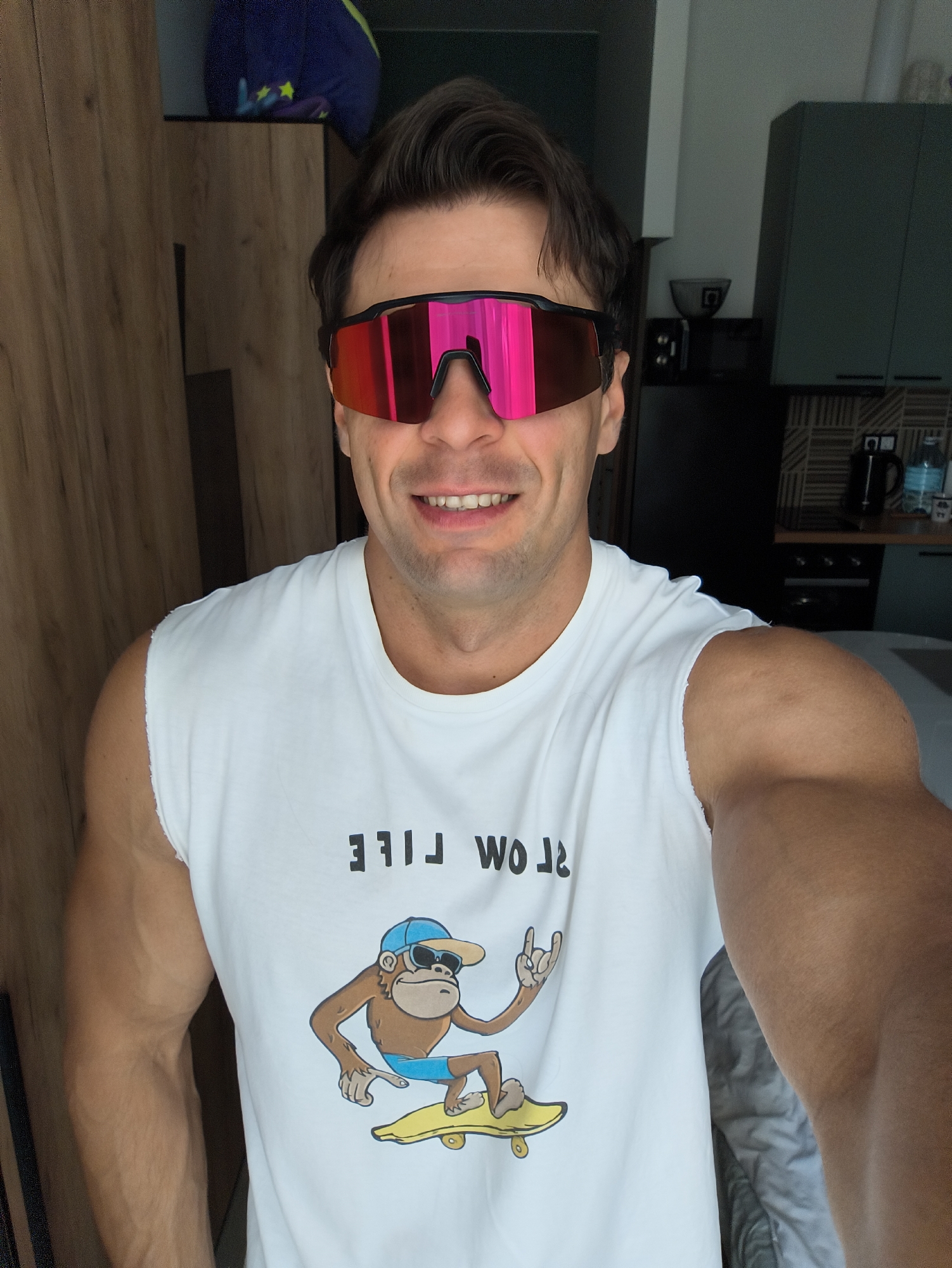 A man is captured in a close-up, waist-up shot, posing in what appears to be an indoor domestic setting, possibly an apartment. The man, with dark hair and a tan complexion, is wearing oversized, bright pink mirrored sunglasses and a white sleeveless t-shirt. The t-shirt features a graphic print of a cartoon monkey wearing a blue baseball cap and sunglasses, riding a yellow skateboard, with the text "WO RILE" printed above it. The man's arms are visibly muscular.
The background suggests a living space with wooden cabinetry on the left and a kitchen area on the right, complete with dark countertops and cabinetry, a kettle, and what looks like a stove. The lighting is bright, indicating it could be daytime. There is no indication of the specific location being Moscow, Russia.