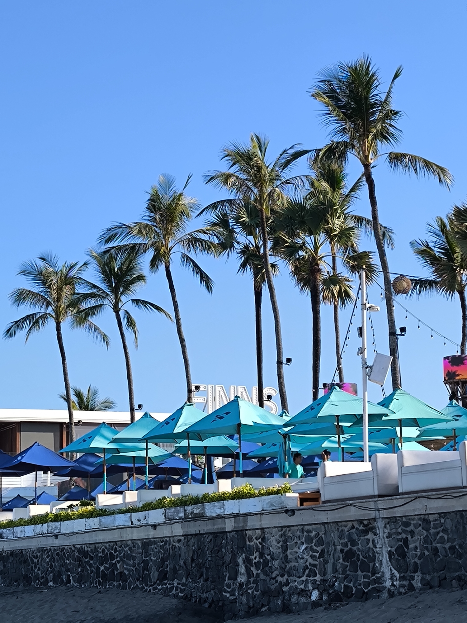 The image depicts a sunny scene at Finns Beach Club in Tibubeneng, Indonesia. Numerous light blue beach umbrellas are arranged in rows along a low wall, interspersed with dark blue umbrellas further back. Palm trees line the scene behind the umbrellas, with a partially visible "FINNS" sign standing behind them. Several people can be seen beneath the umbrellas. In the foreground, there is a stone wall and a glimpse of the beach. A string of lights hangs amongst the palm trees.