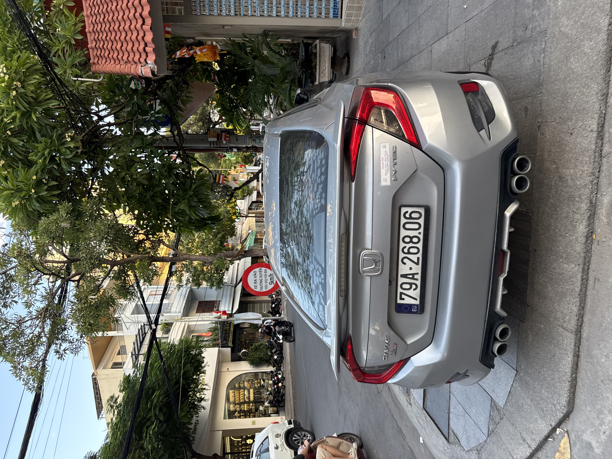 A silver Honda Civic is parked on the side of a street in Nha Trang, Vietnam. The car has a spoiler and dual exhaust pipes. The license plate reads "79A-268.06". There is a red circular sign on a pole behind the car with Vietnamese text. A white SUV is parked in the background. Several motorcycles are also visible on the street. The street is lined with buildings and trees. The sky is blue and clear. The image appears to have been taken during the daytime.