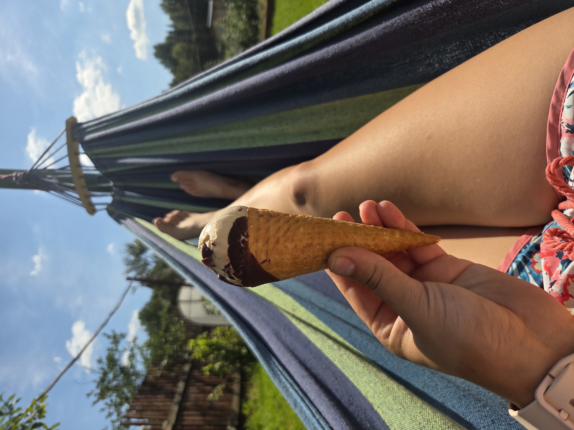 A close-up, first-person perspective shot captures an ice cream cone held in a person's hand, with their legs visible in the background. The person is reclining in a striped hammock, which is predominantly blue, green, and dark purple. The background suggests an outdoor setting with a wooden fence, green grass, trees, and a bright blue sky with scattered clouds. The individual appears to be in a relaxed state, enjoying the ice cream while in the hammock. The image does not provide enough visual cues to determine the specific location within Frolovo, Russia.