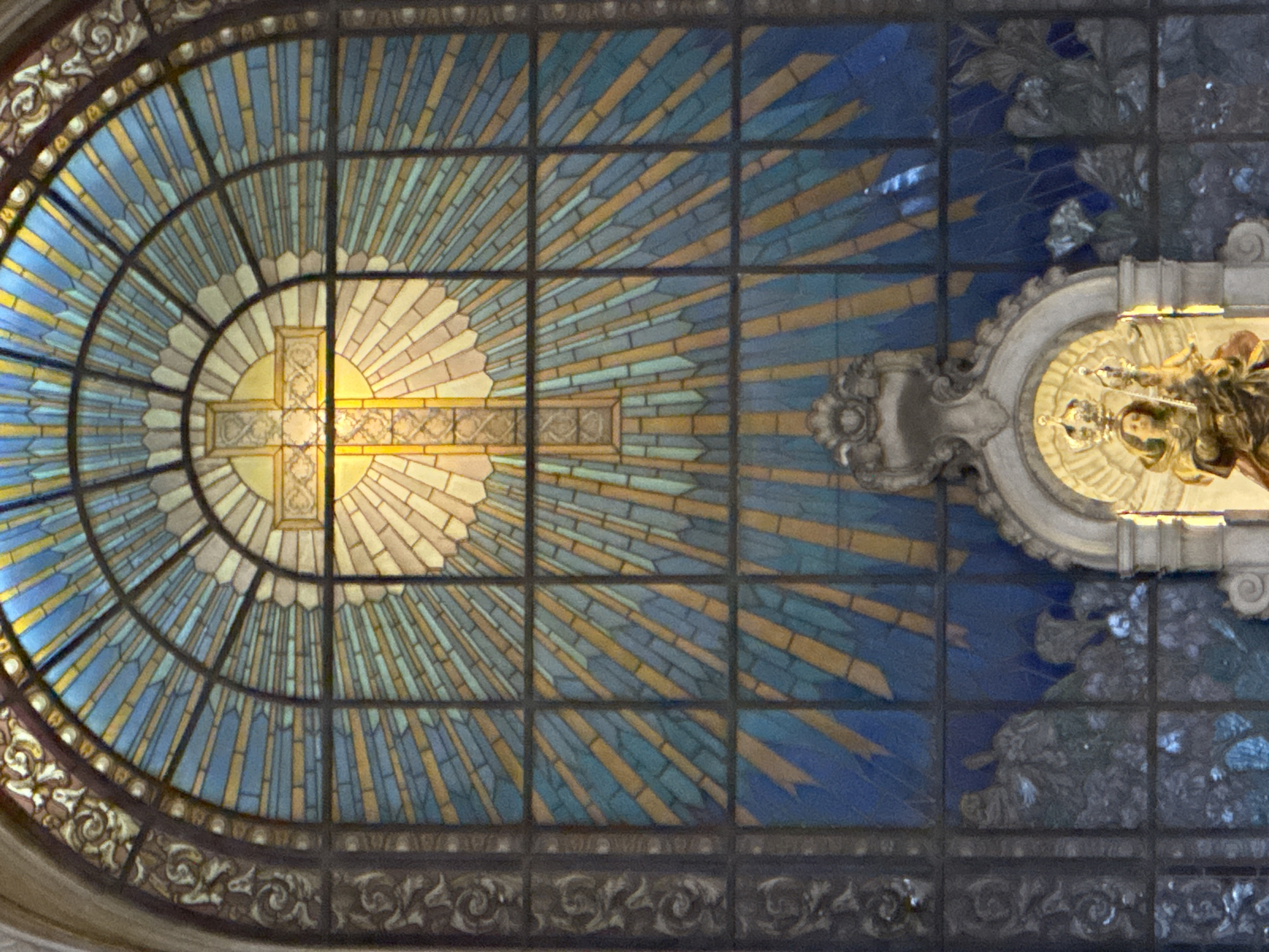 This image shows a stained-glass window above a religious statue in the Church of Our Lady of the Rosary of the Black People (Igreja de Nossa Senhora do Rosário dos Homens Pretos) in Rio de Janeiro, Brazil. The stained-glass window is arched and features a central cross surrounded by radiating lines of yellow and blue glass. The cross itself is depicted with yellow glass and subtle detailing. Below the window is an ornate, light-colored structure framing a statue. The statue appears to be of a crowned woman holding a child, both with golden halos and elaborate robes. The scene is indoors, with the lighting suggesting it is daytime, illuminating the stained glass from behind. The lower portion of the image shows darker, possibly more abstract, stained glass depicting figures or scenes in deep blues and greys. The overall impression is one of religious art and architecture within a church setting.