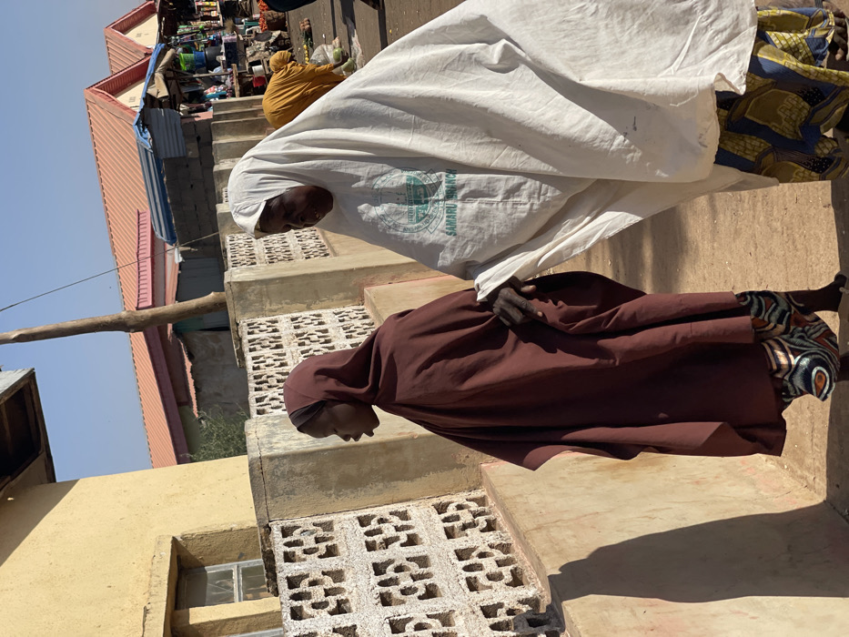 The image displays an outdoor scene in Samaru, Nigeria, under bright daylight. In the foreground, two women walk from left to right. The woman on the left, positioned in profile facing left with her mouth slightly open, wears a dark reddish-brown head covering and a loose matching garment over patterned trousers. She holds the right forearm of the woman to her right. This second woman, facing slightly towards the viewer, wears a white head covering and a loose white garment featuring a circular green emblem inscribed with "SAMARU BRANCH." She also wears patterned trousers.
To the immediate left, a low wall constructed from patterned concrete blocks runs parallel to a light yellow-ochre building with a single window. In the mid-ground, a tall wooden pole stands vertically. The background reveals a bustling market environment with multiple stalls situated under structures topped with orange-red corrugated metal roofs. Various goods are displayed on the stalls, and several other individuals, including one person in a yellow garment bending over a stall, are visible further back. The ground appears to be unpaved dirt, and strong sunlight casts distinct shadows across the scene.