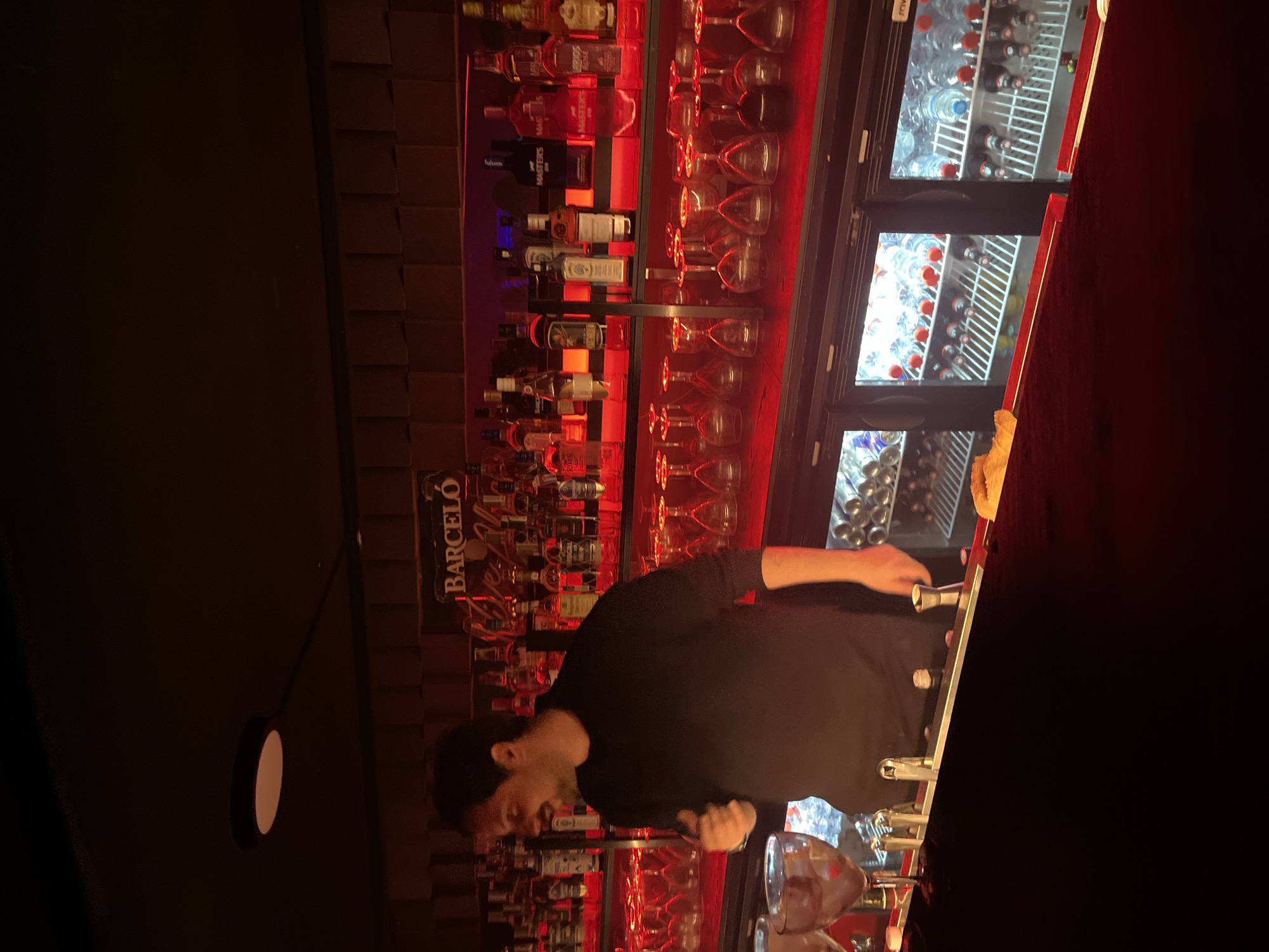 The image depicts a bartender working at a Barceló-branded bar. Bottles of alcohol and rows of clean glasses are displayed on shelves behind the bar, illuminated by red lighting. There is a refrigerator behind the bar stocked with bottled drinks. The bartender, dressed in black, is standing behind the bar with his hands near the counter. The location is likely Valencia, Spain, due to the "Barceló" sign.