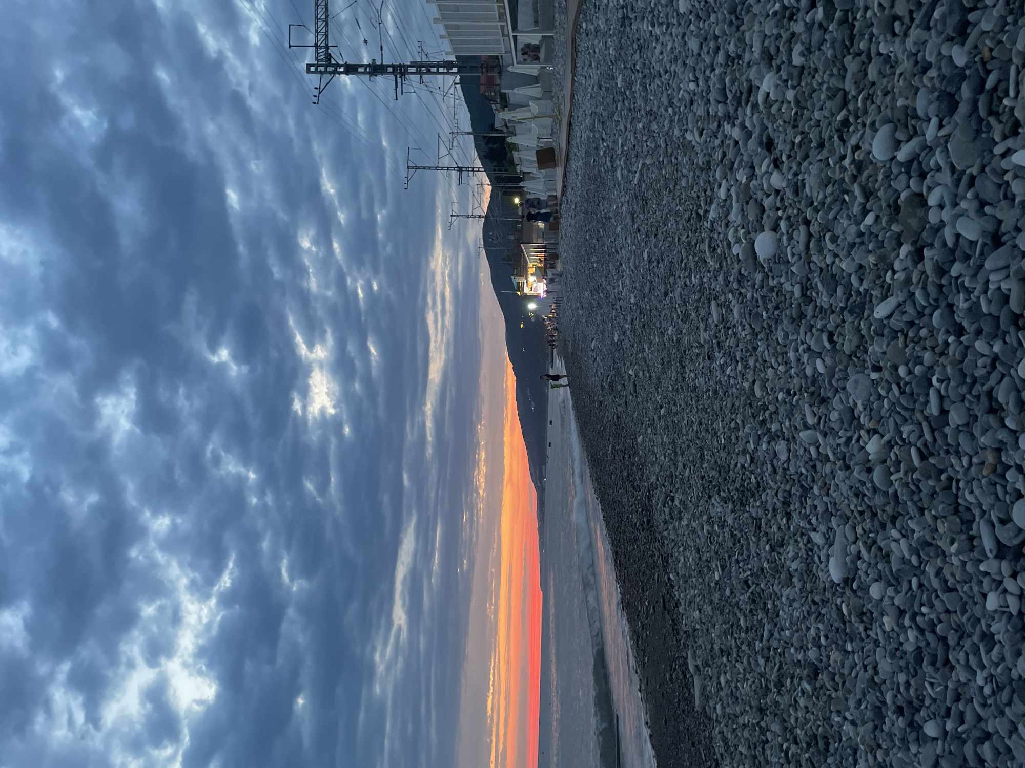 The image captures a view of a pebble beach at dusk. The sky is filled with dramatic, dark gray clouds with patches of lighter sky and sunset colors, ranging from orange to red, visible on the horizon over the sea. The sea is calm with small waves rolling onto the shore. The beach is densely covered with smooth, grey pebbles. To the right of the beach, a promenade or walkway is visible, lined with white lounge chairs and umbrellas. Beyond that, there are buildings with bright lights, suggesting a commercial area or cafes. Overhead, there are numerous electrical poles and wires, indicating proximity to a railway line. A few people are visible walking along the beach and the promenade. Mountains form a backdrop in the distance, silhouetted against the twilight sky. The overall atmosphere is serene and dusky.