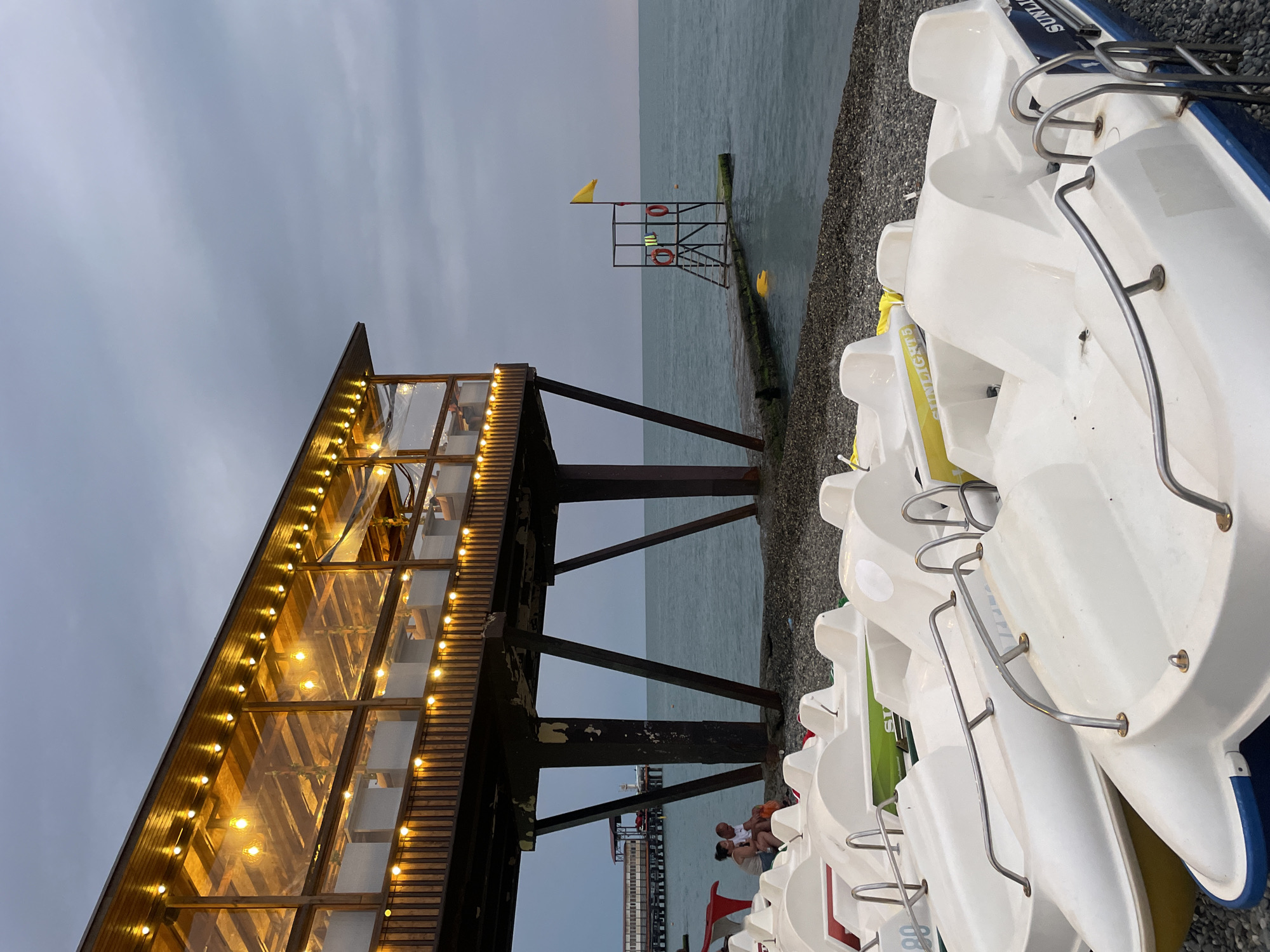 At an outdoor cafe on the coast of Sochi, Russia, a couple sits talking under the shelter of a wooden structure illuminated by warm string lights. The cafe extends over a pebbled beach lined with white pedal boats. In the distance, the calm sea stretches to the horizon under a cloudy sky. A lifeguard tower stands on a pier, adorned with a yellow flag. The overall atmosphere is serene and peaceful, suggesting a late afternoon or early evening scene.