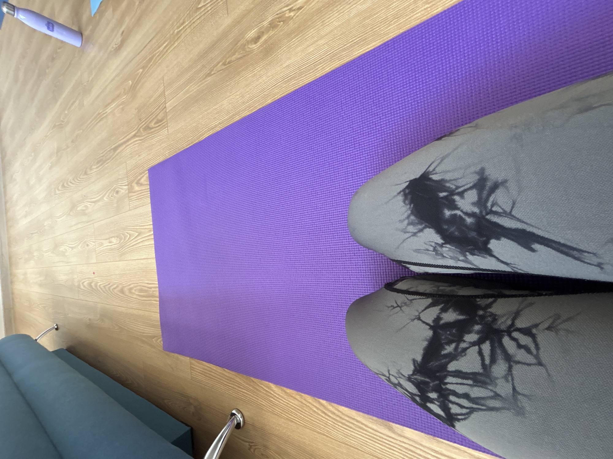 A top-down shot shows a purple yoga mat on a light wood-toned floor, with a blue sofa at the lower left and a light purple water bottle at the upper left. Two knees covered in grey leggings with a black abstract pattern are visible on the right of the yoga mat. No people are definitively seen. The scene likely takes place indoors, given the furnishings and floor. Location information, Sofia, Bulgaria is only presumed and not visible from the image alone.